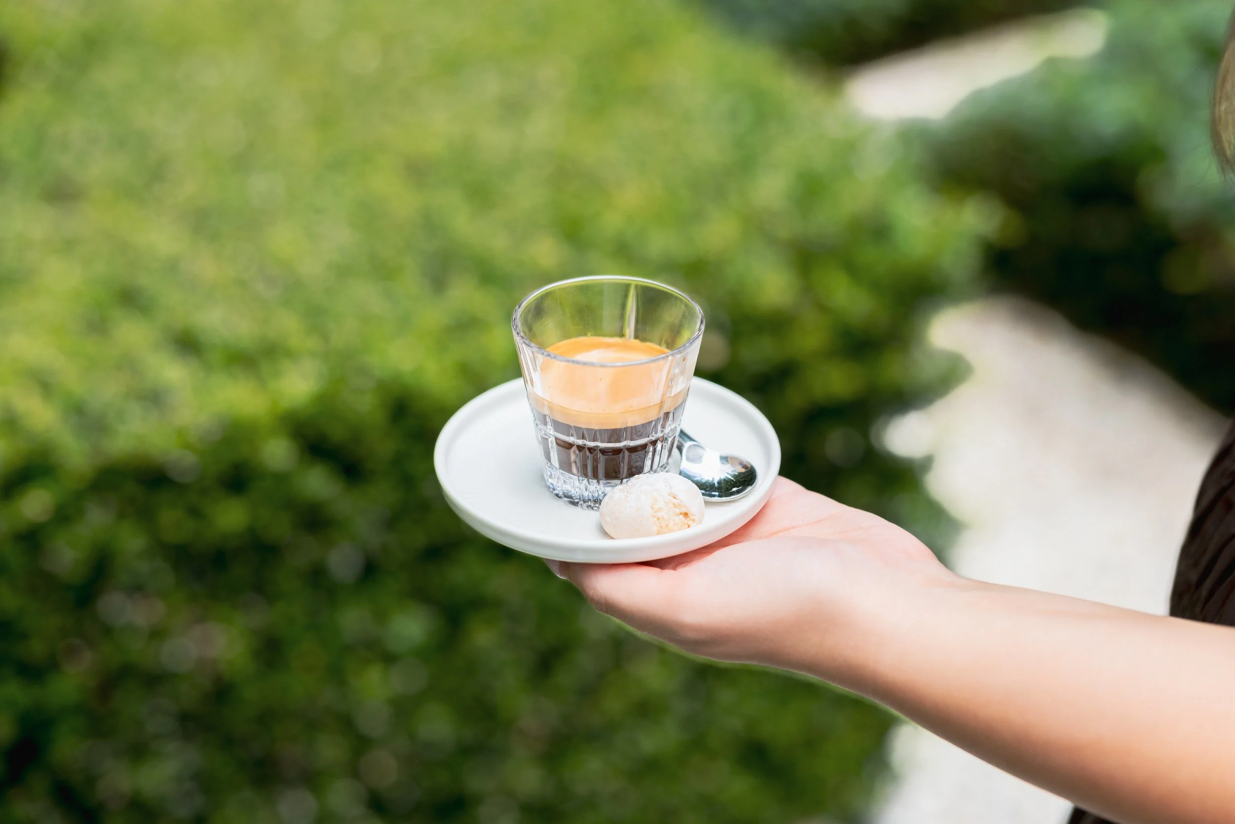A person holding a white plate with a glass of espresso, a spoon, and a small cookie against a blurred green outdoor background.