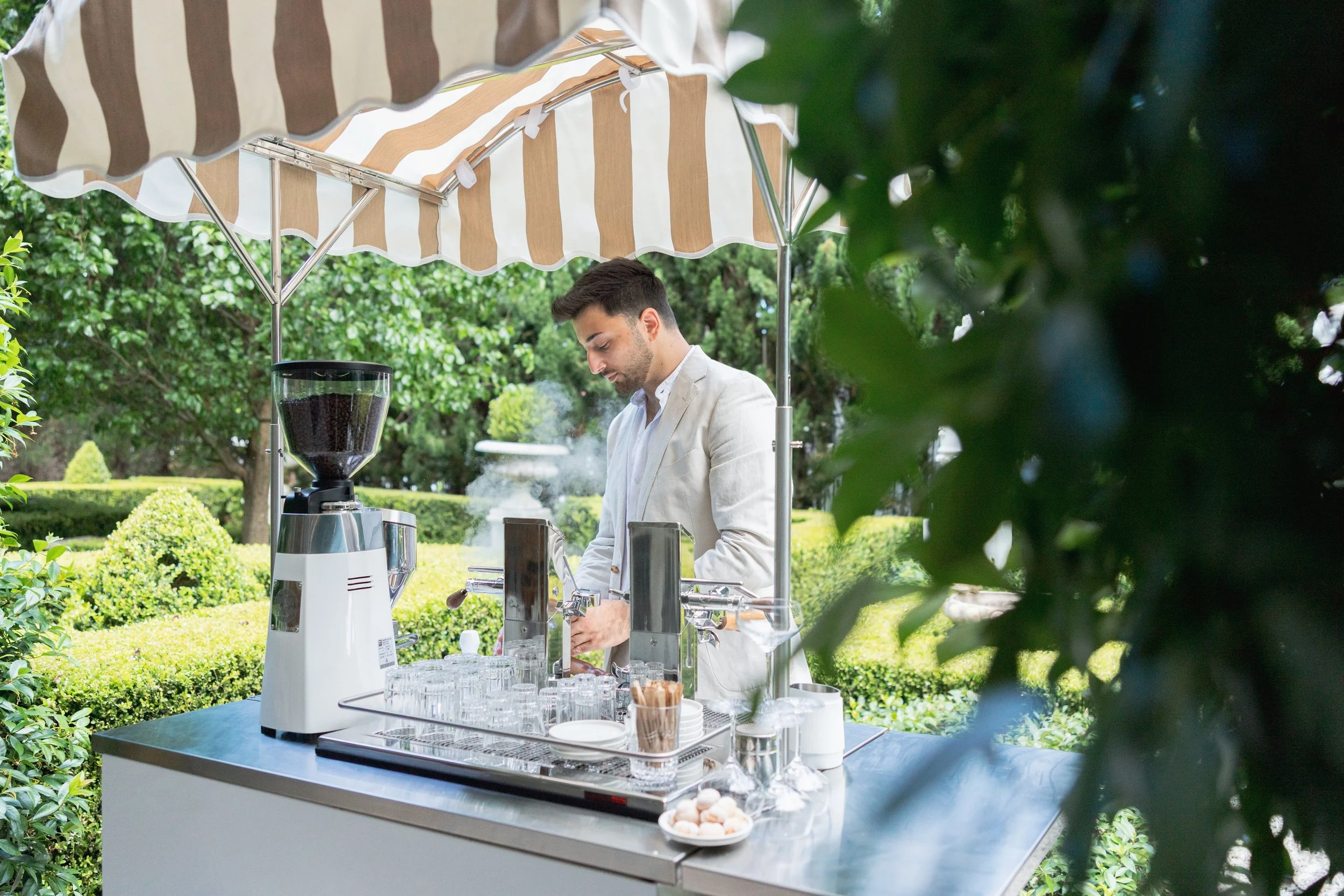 A man in a white suit making coffee at an outdoor coffee stand under a brown and white striped umbrella, surrounded by lush green bushes and trees.