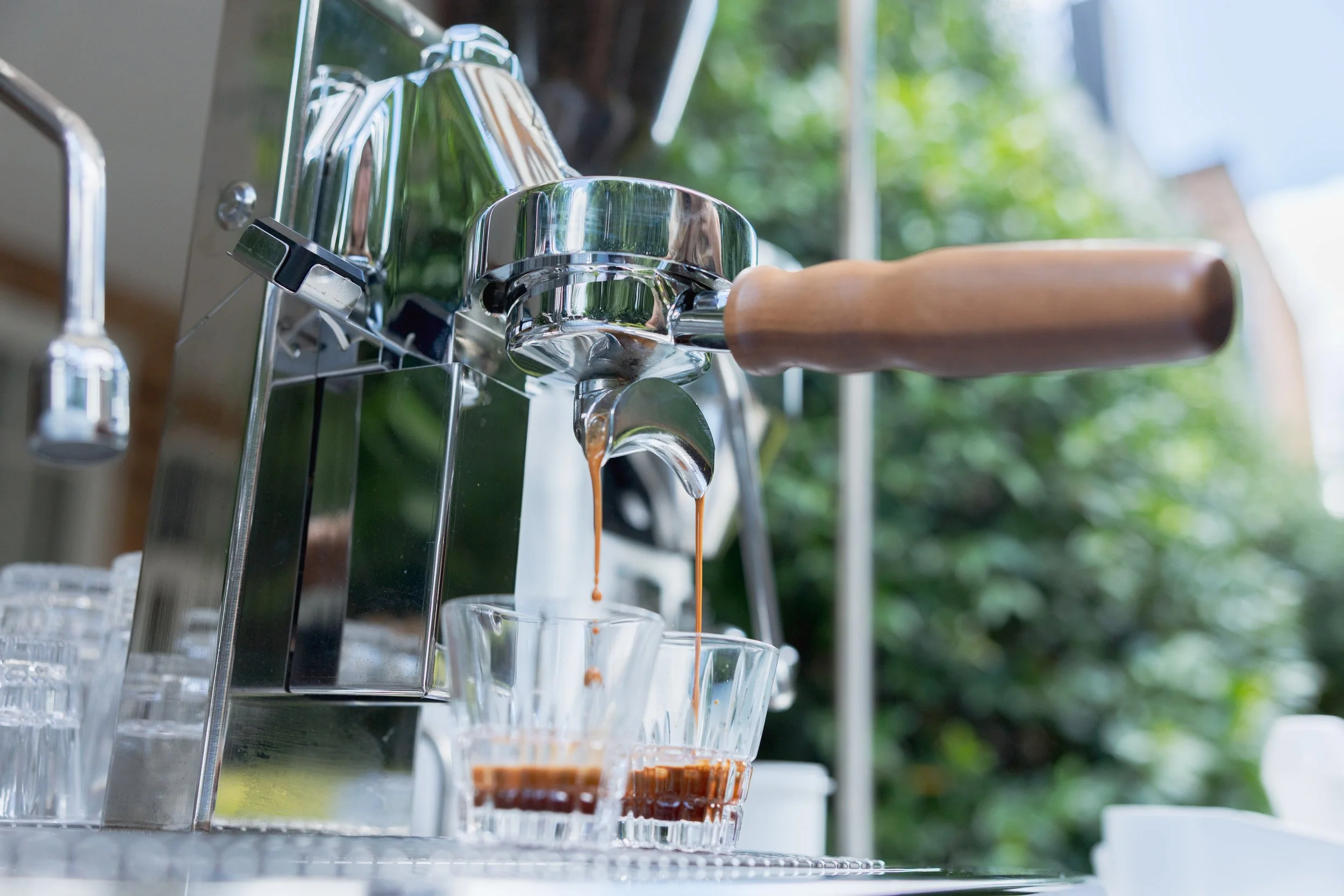 An espresso machine brewing coffee into two clear glasses with wood handles, with a lush green outdoor background.