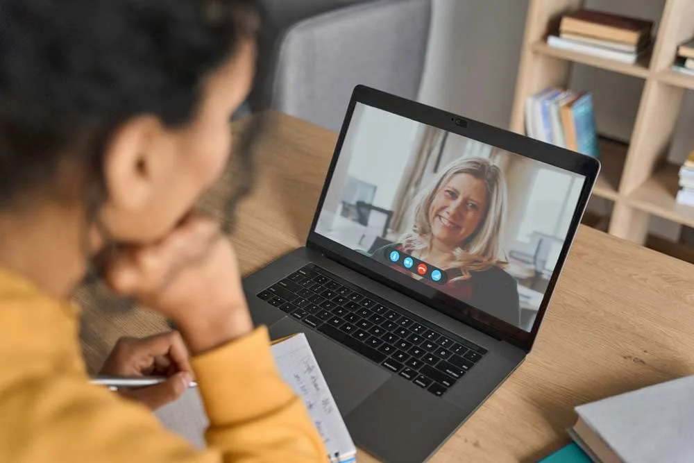 A person with glasses and curly hair taking notes while on a video call with Keri Schwebius on a laptop screen in a home office setting, with bookshelves in the background.
