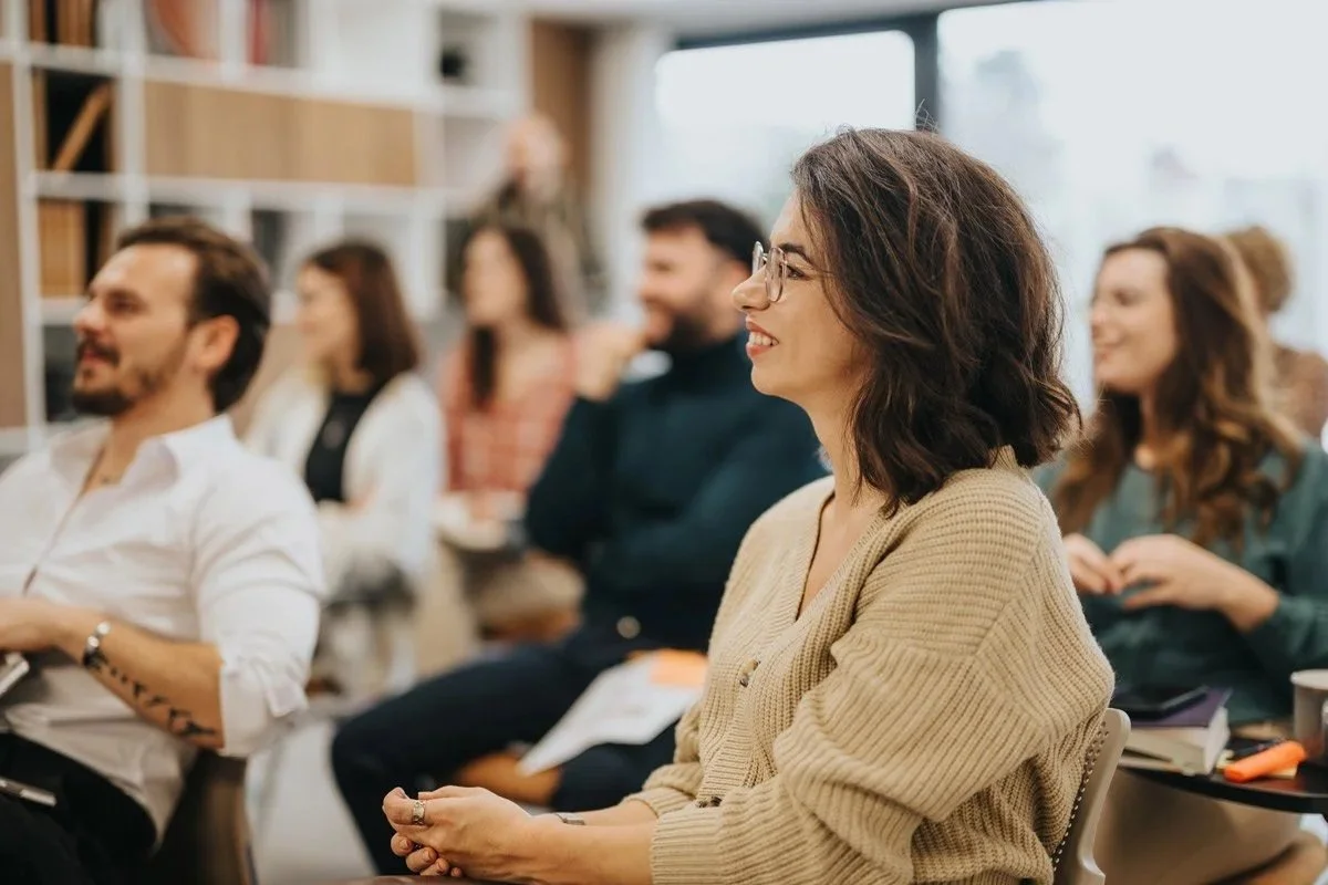 People attending a seminar or workshop, sitting and listening attentively, with some taking notes, in an indoor setting with large windows.