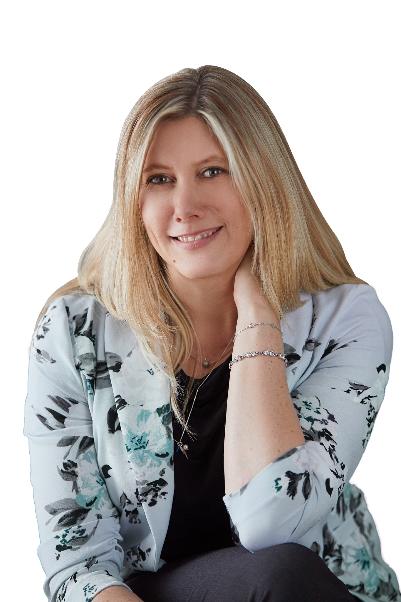 A woman with blonde hair smiling, wearing a light-colored floral blazer, sitting with her hand resting on her neck.