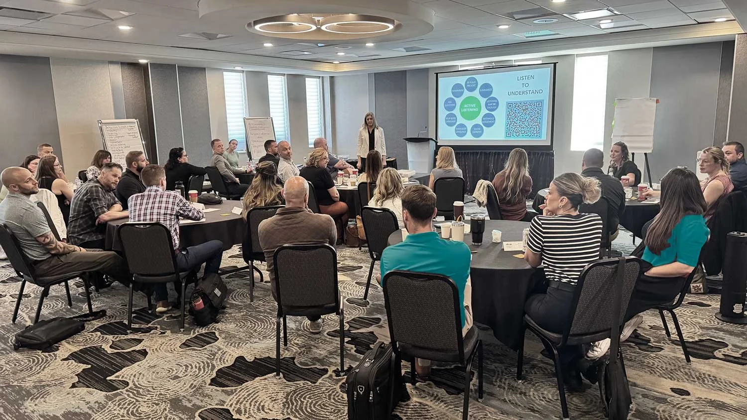 A large group of people attending a conference or seminar in a spacious hotel meeting room, seated at round tables, listening to a speaker at a podium with a slide presentation projected on a screen behind her.