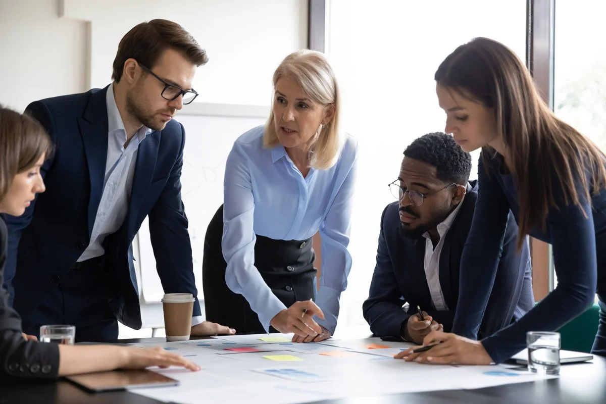 A diverse group of five professionals gathered around a table in a business meeting, actively engaged in a discussion while looking at documents and charts.