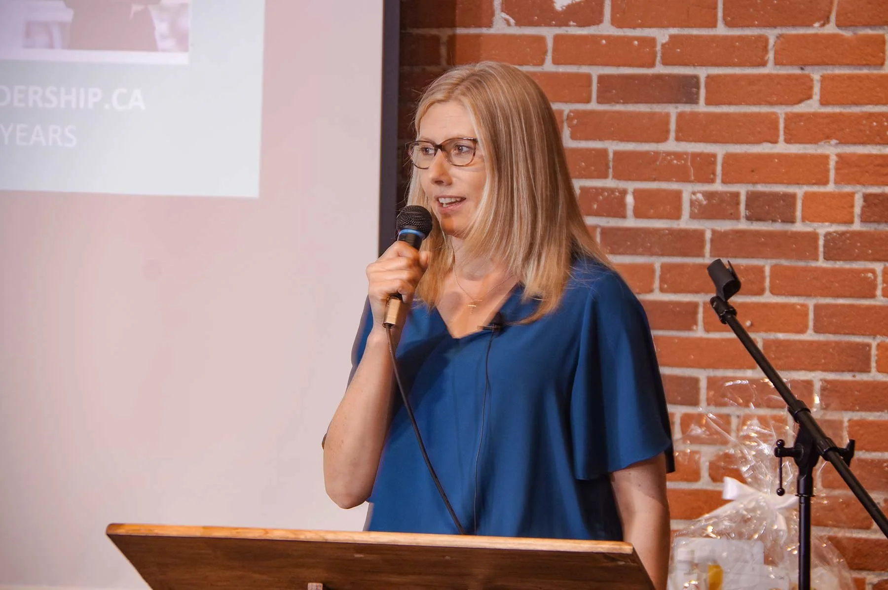 Keri Schwebius speaking into a microphone at a podium, with a brick wall and a projection screen in the background.
