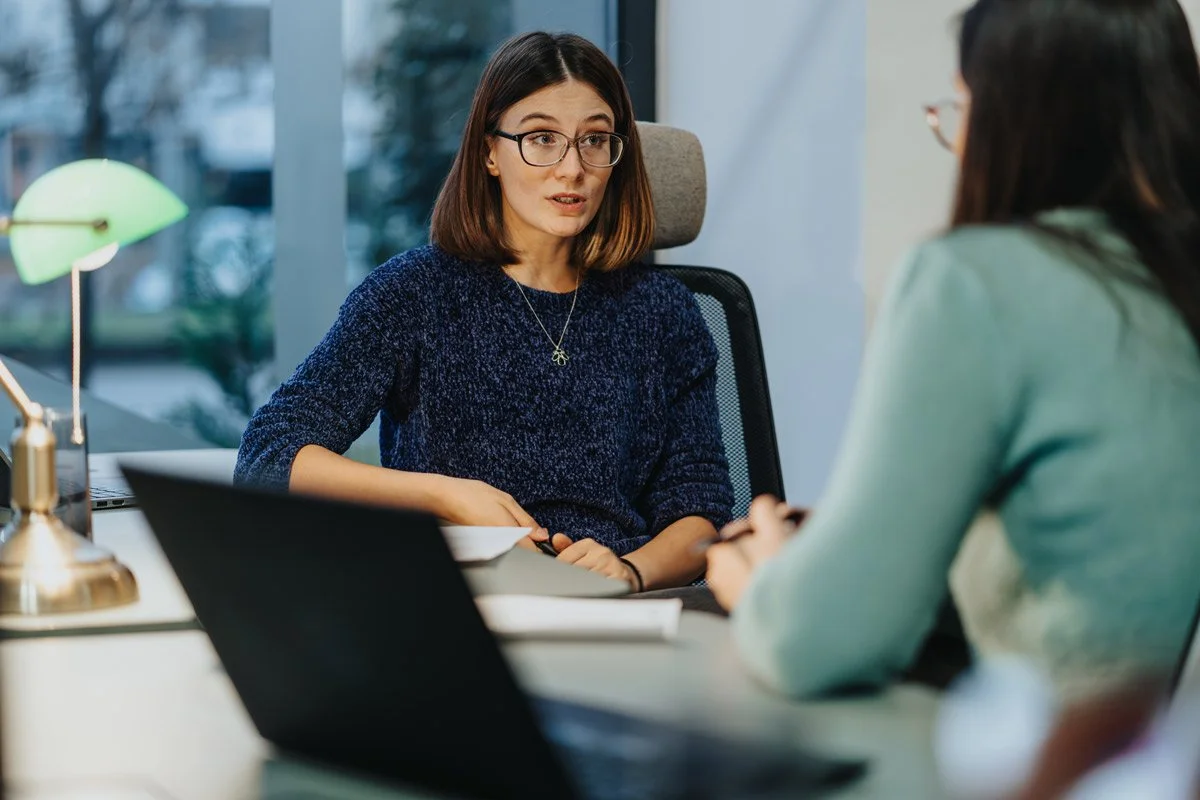 Two women having a conversation in an office, one wearing glasses and a blue sweater, the other with dark hair and a light sweater