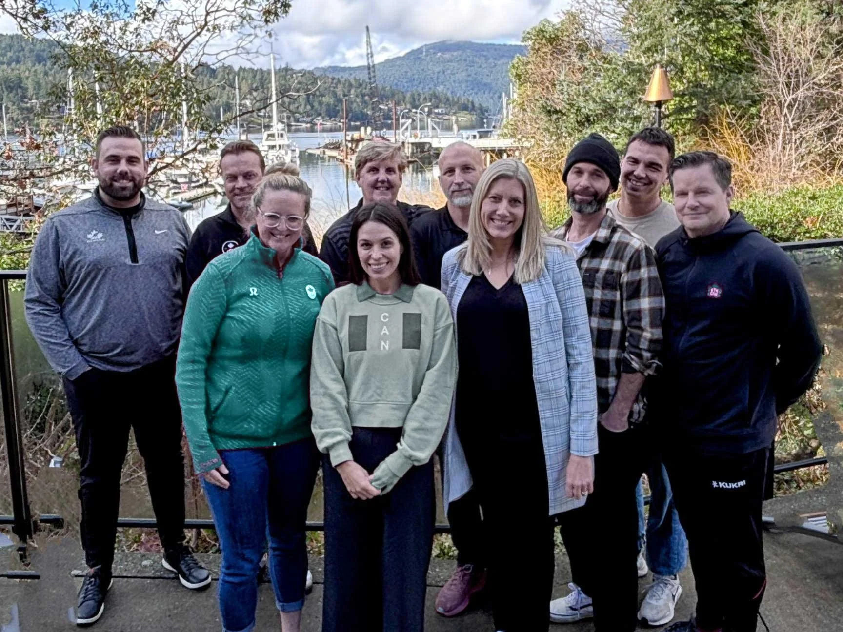 Keri Schwebius with a group of eleven people, mixed genders, standing outdoors near a water body with boats, trees, and mountains in the background, posing for a group photo for Ellevate Team Training.