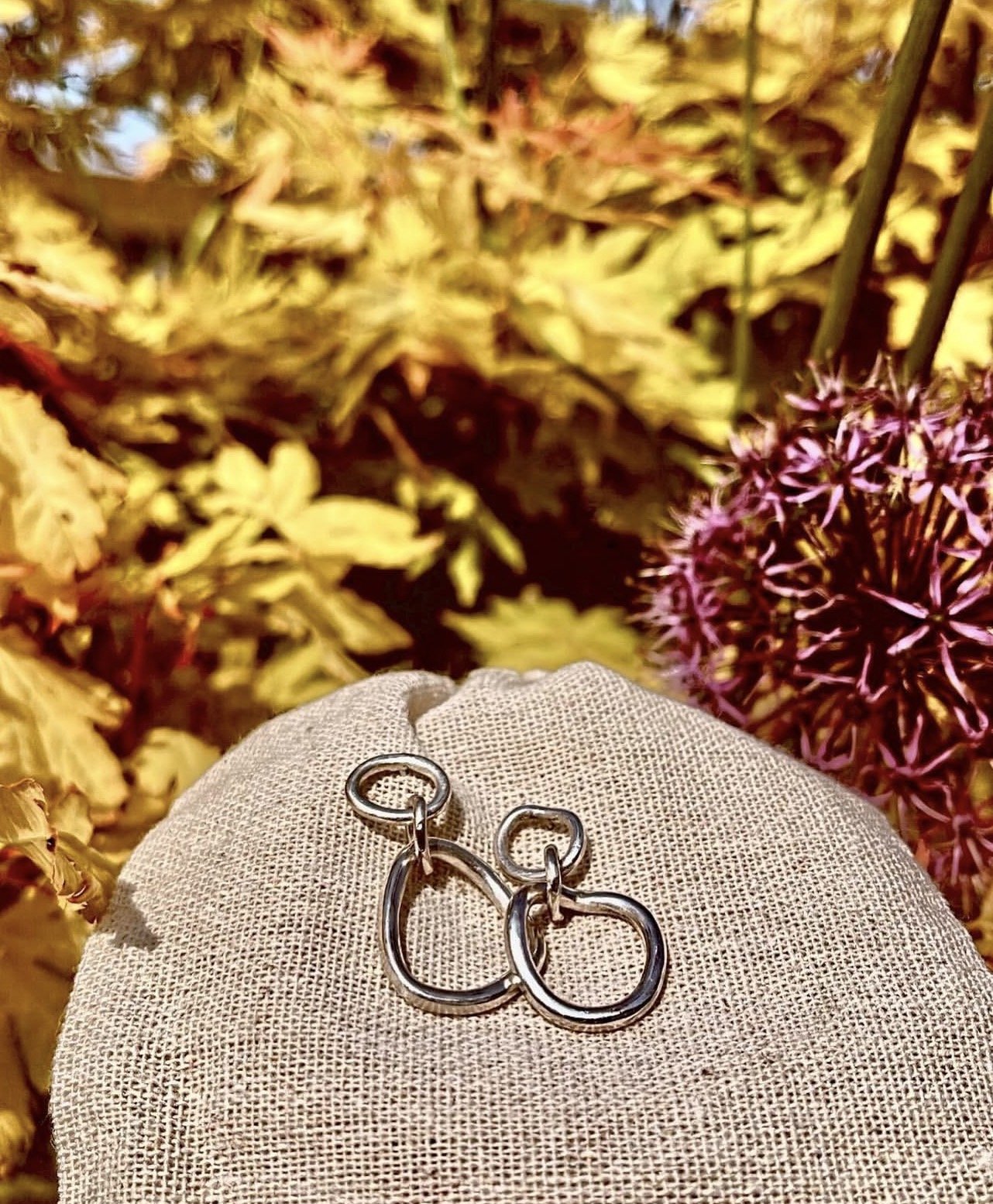 A pair of silver hoop earrings on a beige fabric surface with dried yellow leaves and purple flowers in the background.