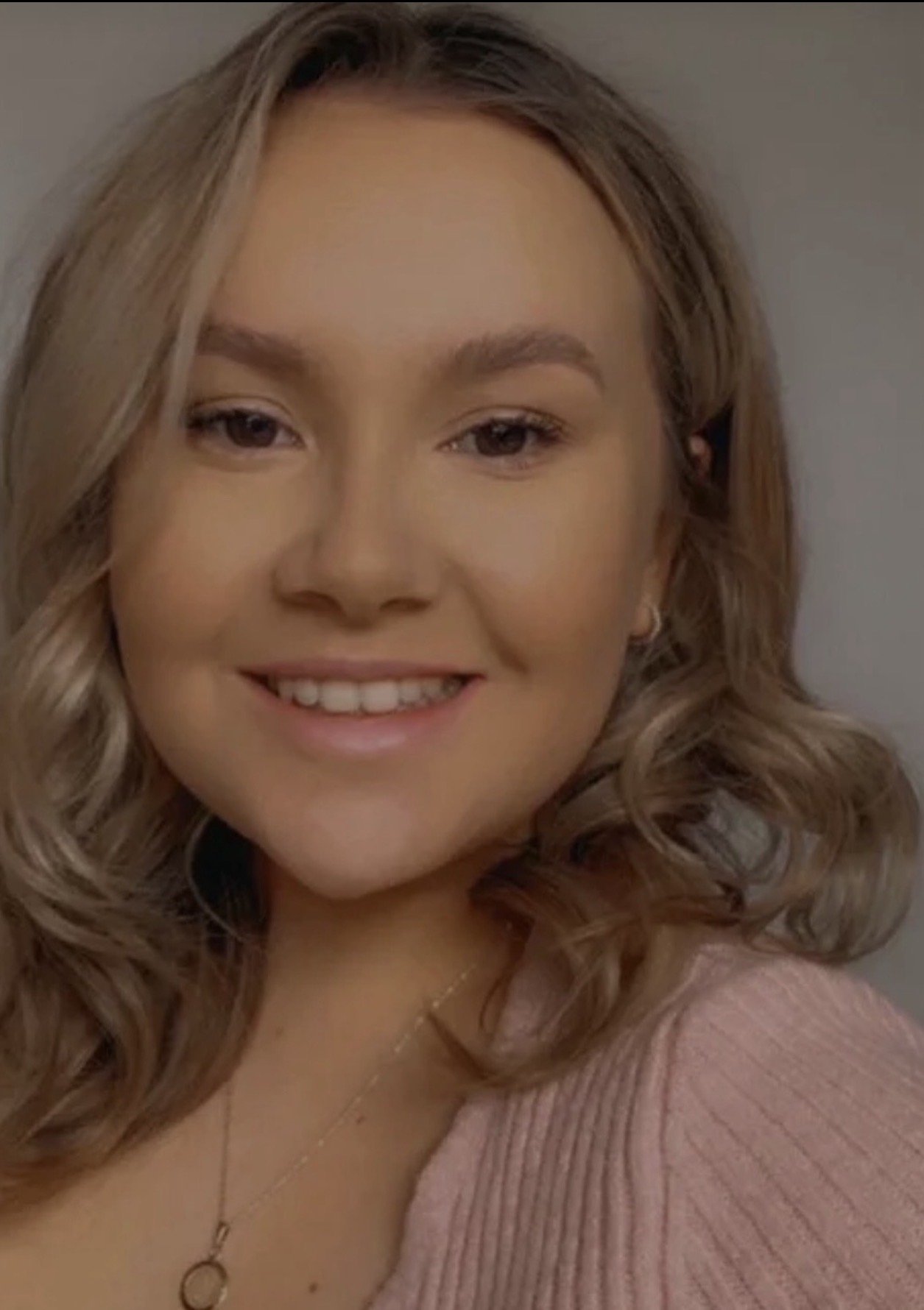 Close-up of a smiling young woman with wavy, shoulder-length blonde hair, wearing a pink top and a delicate necklace.