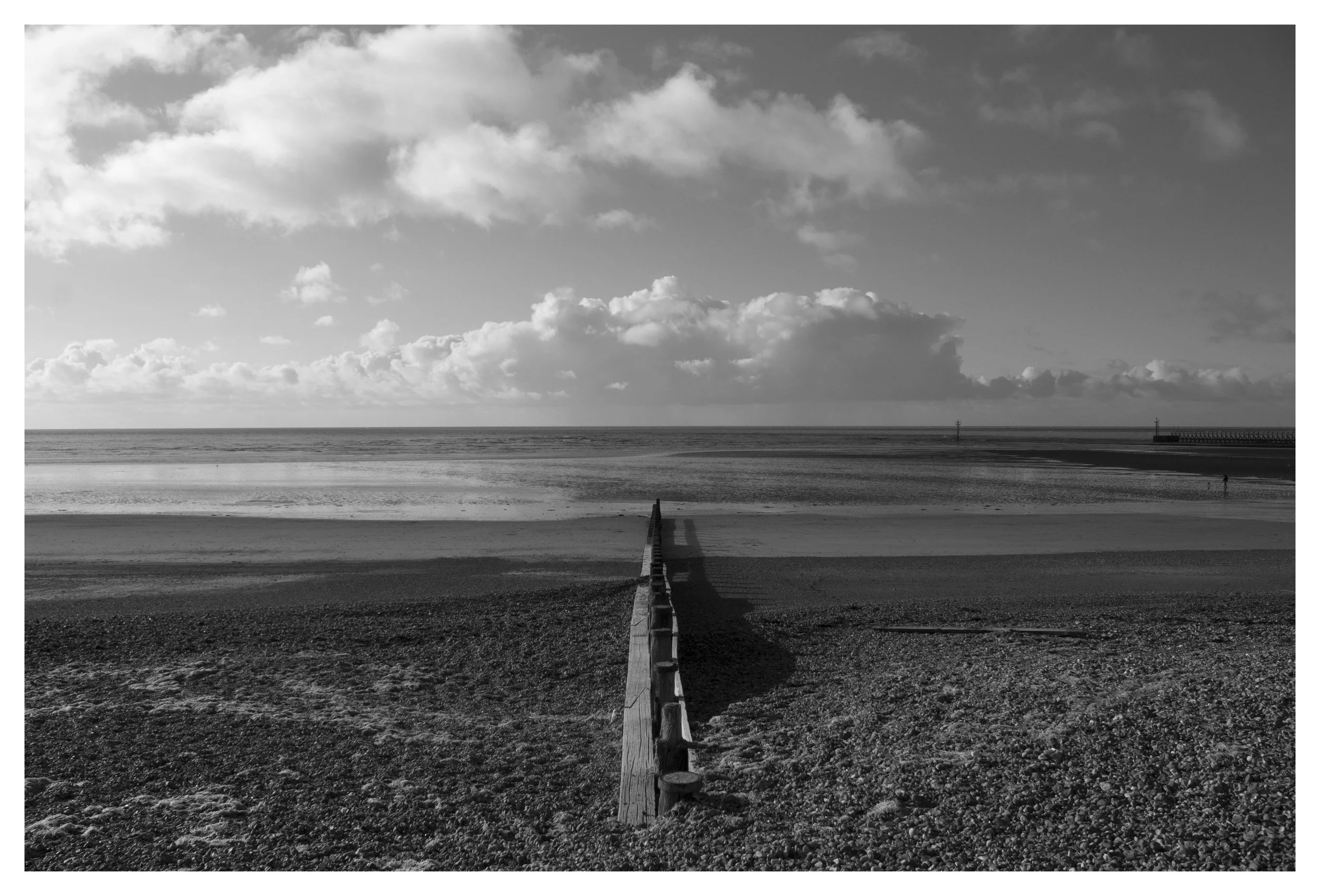 Black and white photo of a beach with a wooden barrier leading to the ocean, cloudy sky in the background, and a pier on the right side.