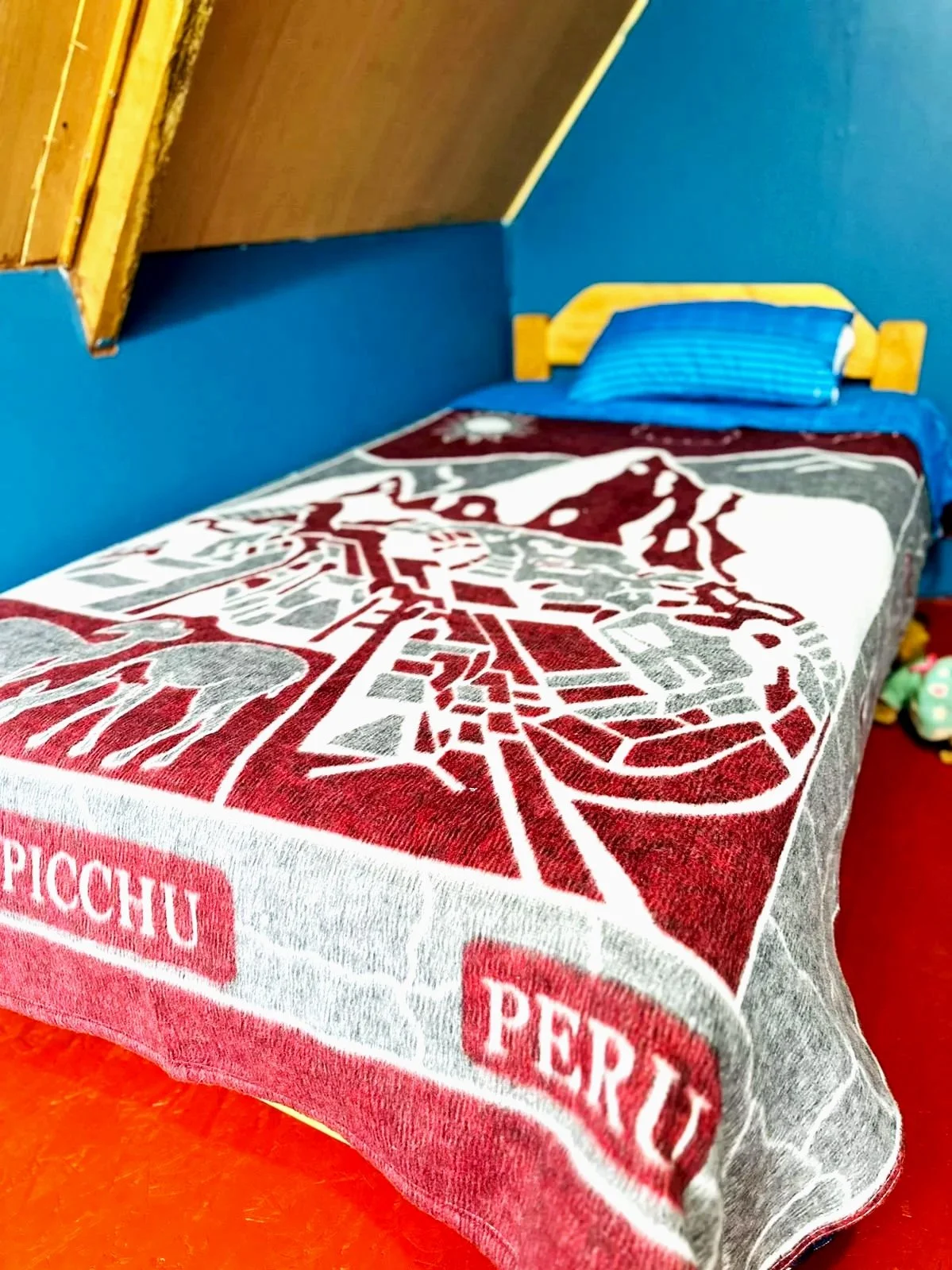 A bed with a decorative blanket featuring a red and gray design of the Piazza del Duomo in Perugia, Italy, and a blue pillow with yellow accents in a small bedroom with blue walls and wooden furniture.