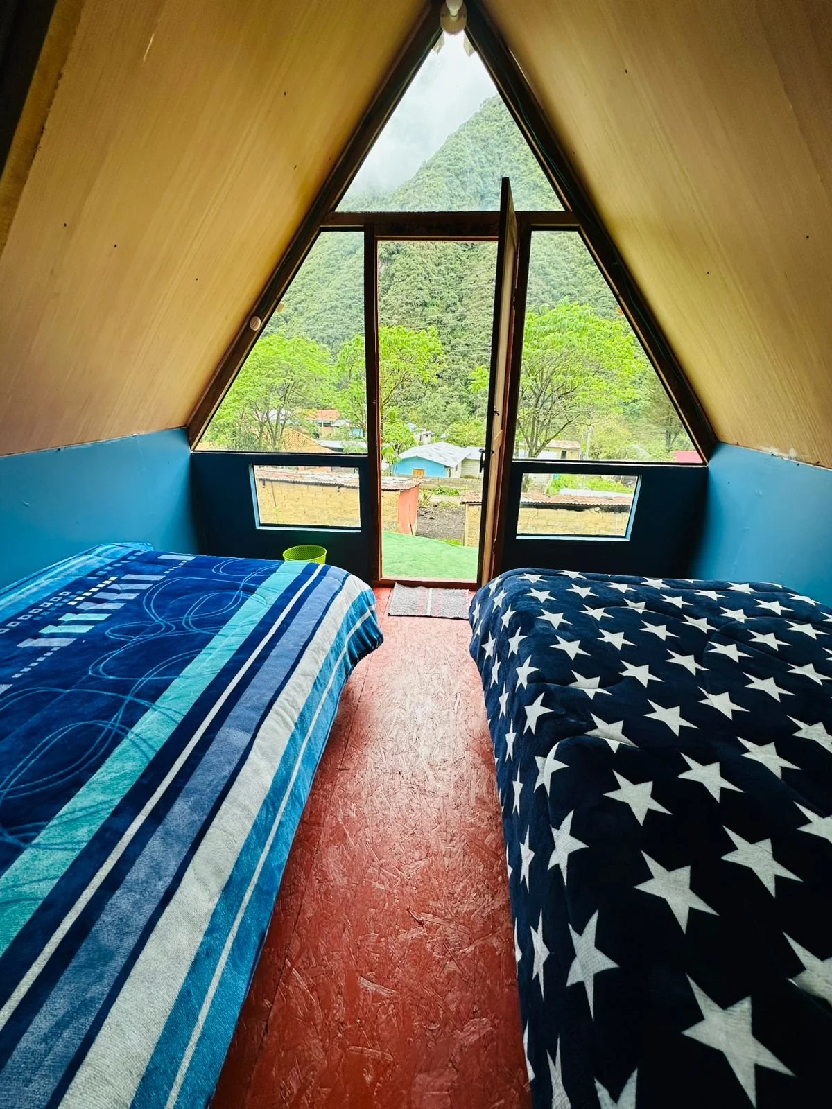 Interior of a triangular-shaped room with two beds, one with a blue striped bedspread and the other with a star pattern, facing large triangular windows showing mountains and greenery outside.