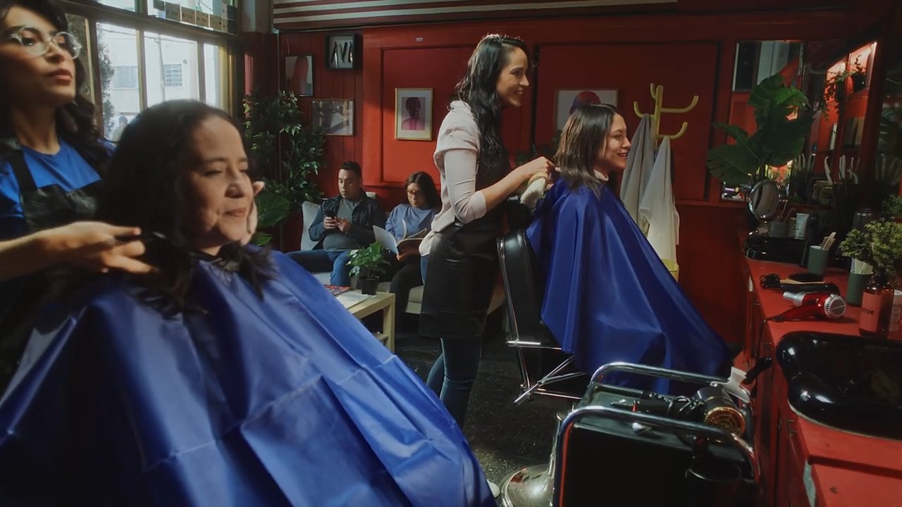 Women getting haircuts at a salon with stylists and customers, blue capes, and salon equipment.