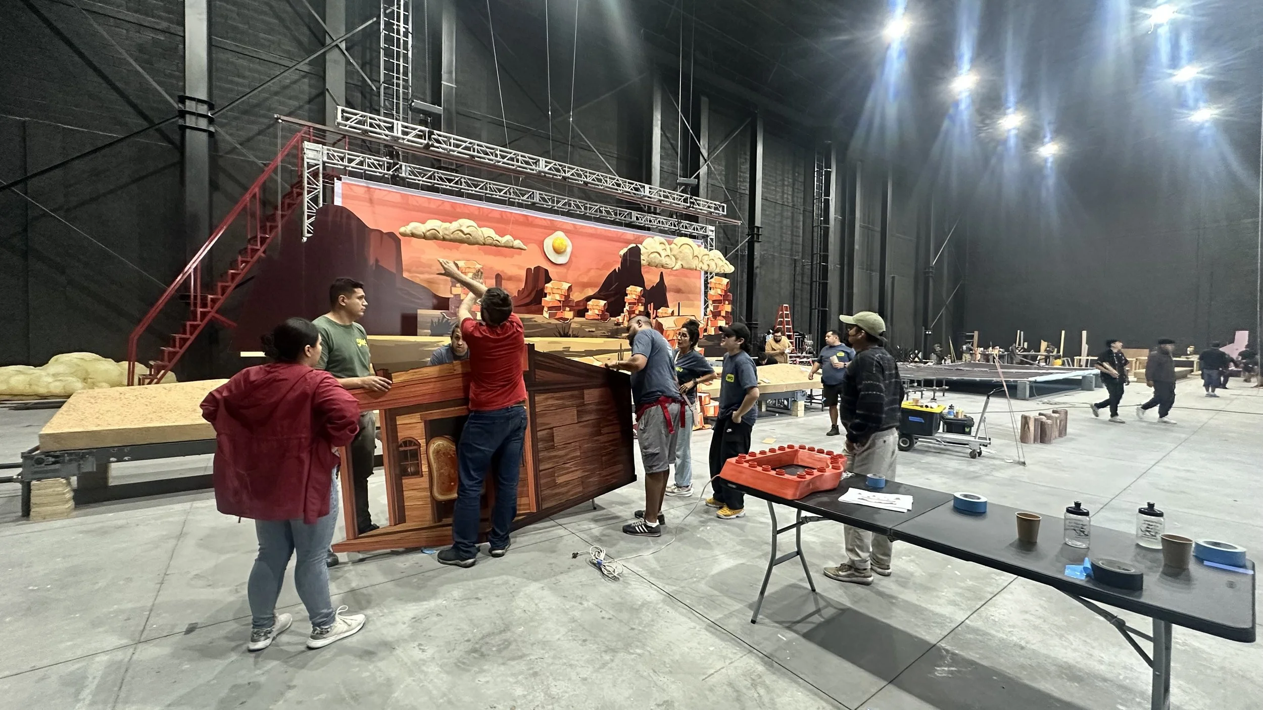 Group of people setting up a stage for a theatrical performance in a large indoor space. The background features a painted scenic backdrop of desert mountains with a sunset and clouds, and props resembling a desert town.