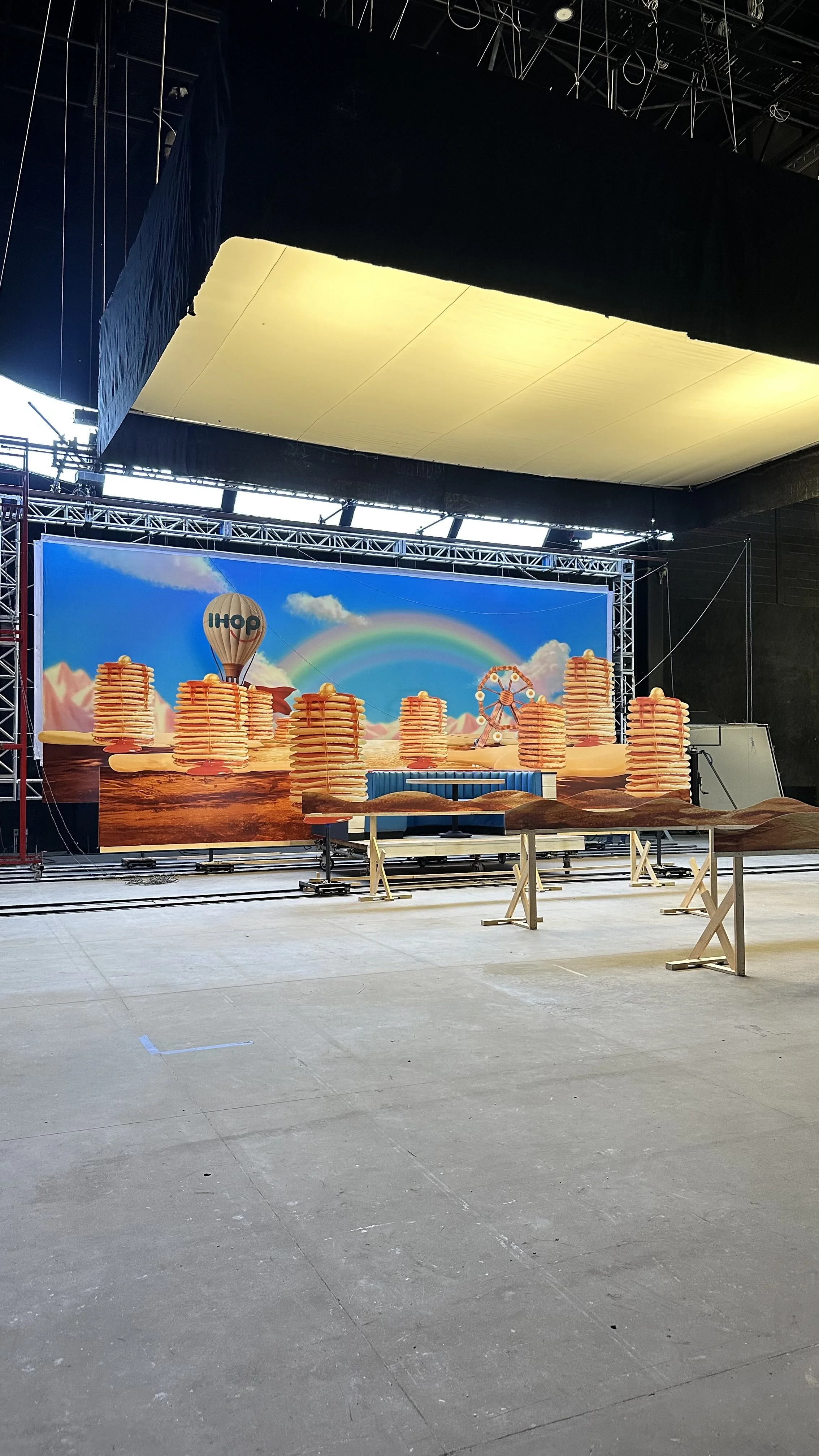 Stage with a colorful backdrop showing stacked pancakes, a rainbow, a hot air balloon with 'IHOP' logo, and a Ferris wheel, with scaffolding and tables in the foreground, likely preparing for an IHOP promotional event or performance.