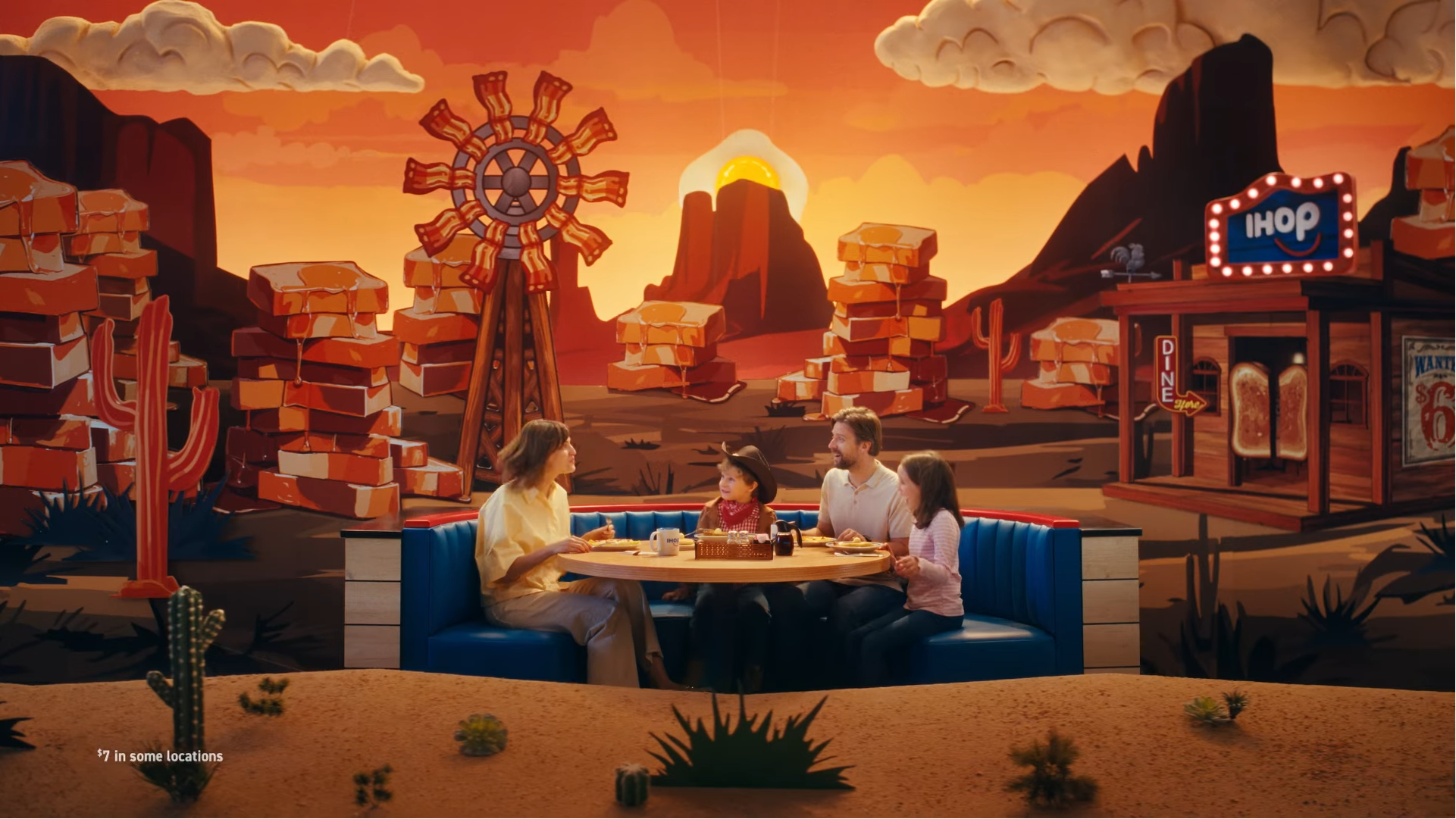 Family of four sitting at a booth in a themed restaurant with a desert scene mural, featuring a windmill, cacti, mountains, and a sunset.
