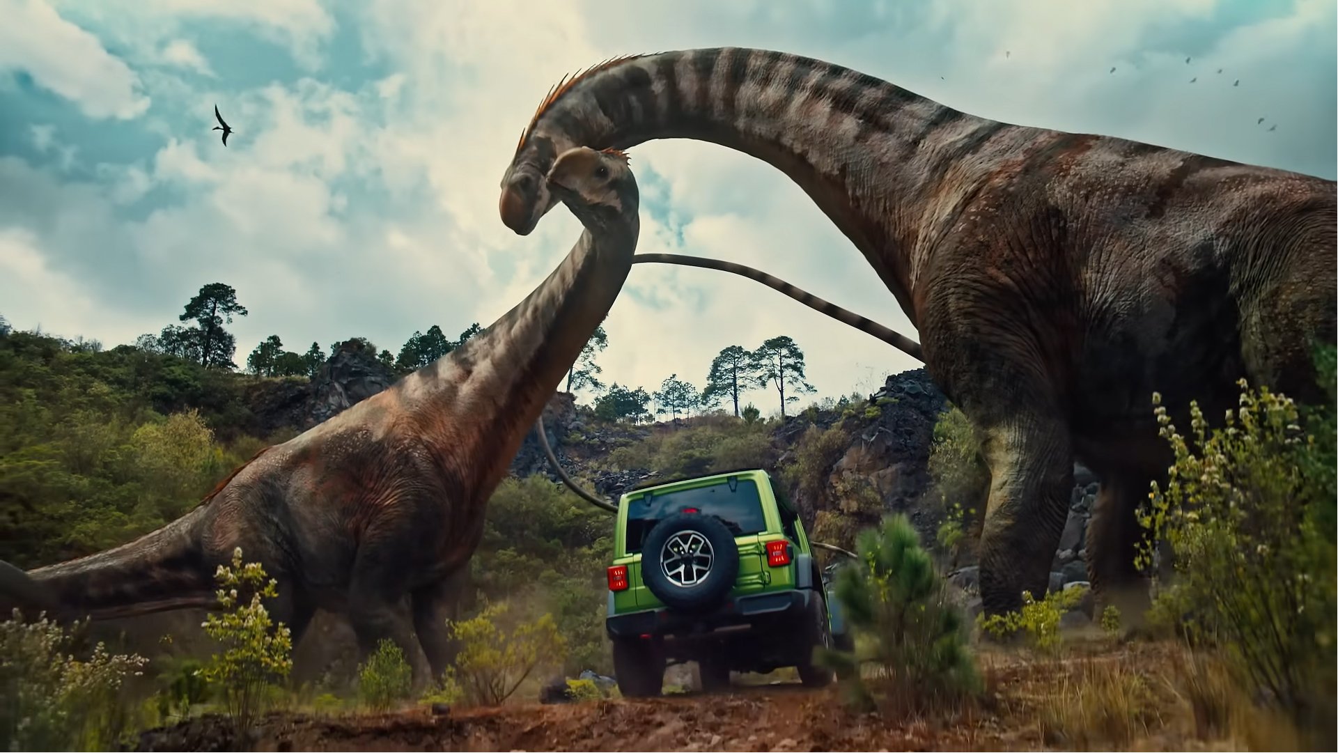 Two dinosaurs, one large with a long neck and the other smaller, are fighting in a prehistoric landscape with trees and mountains in the background. There is a green off-road vehicle with a spare tire mounted on the back in the scene.