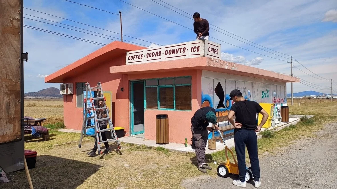 People outside a small pink building with a sign offering coffee, sodas, donuts, ice, chips, while one person on the roof repairs or paints it, and two others work near the entrance, with a scenic rural landscape and blue sky in the background.