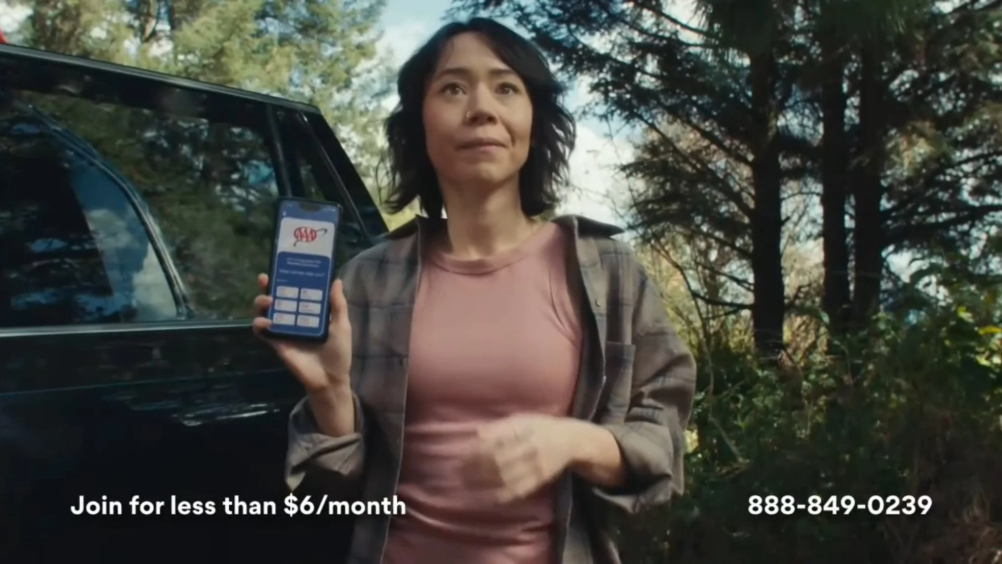 A woman standing outdoors near a black car, holding a smartphone displaying a login screen with a red logo, surrounded by trees and foliage.