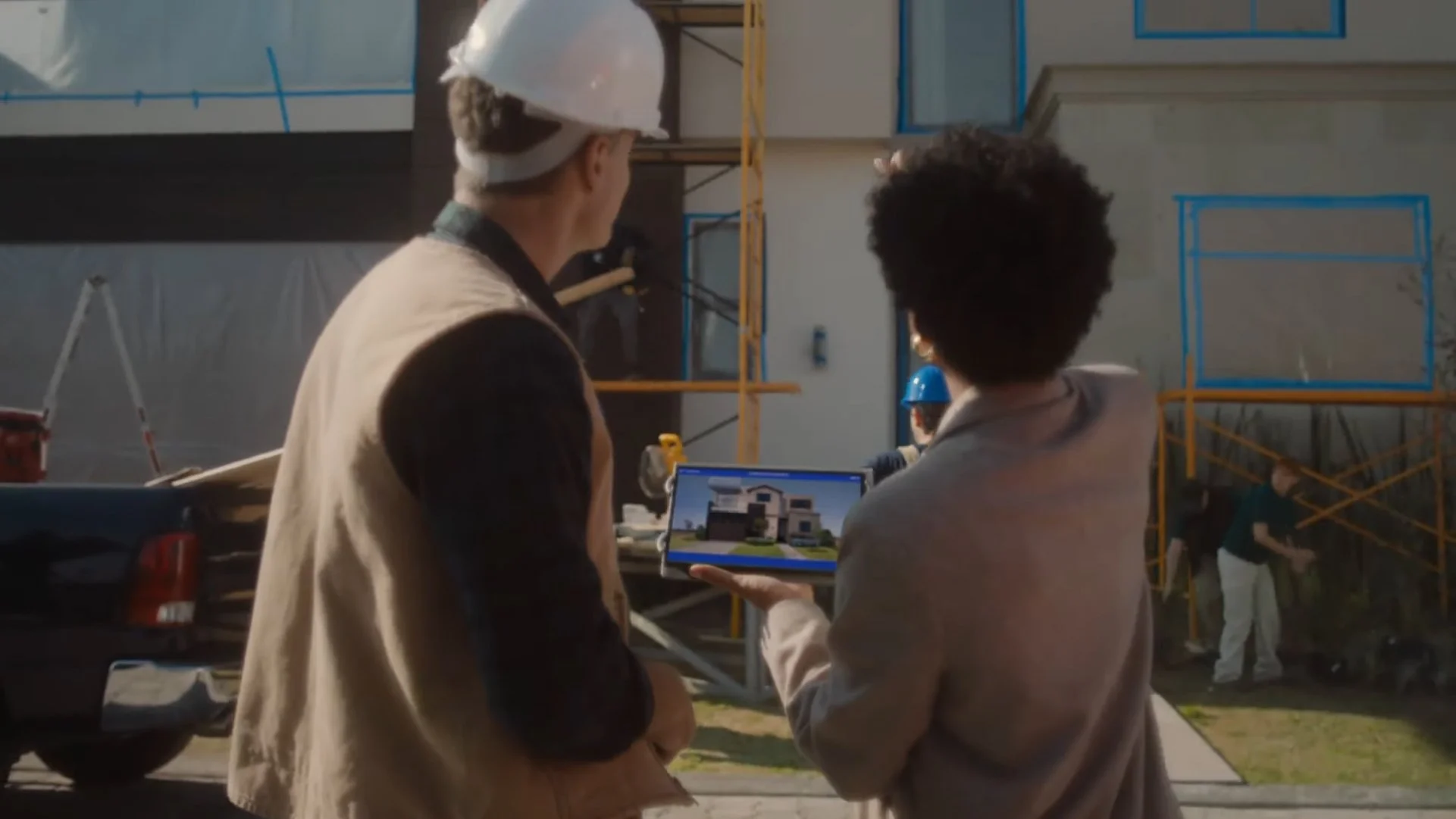 Two people are looking at a tablet with a house plan on a construction site, with scaffolding and workers in the background.
