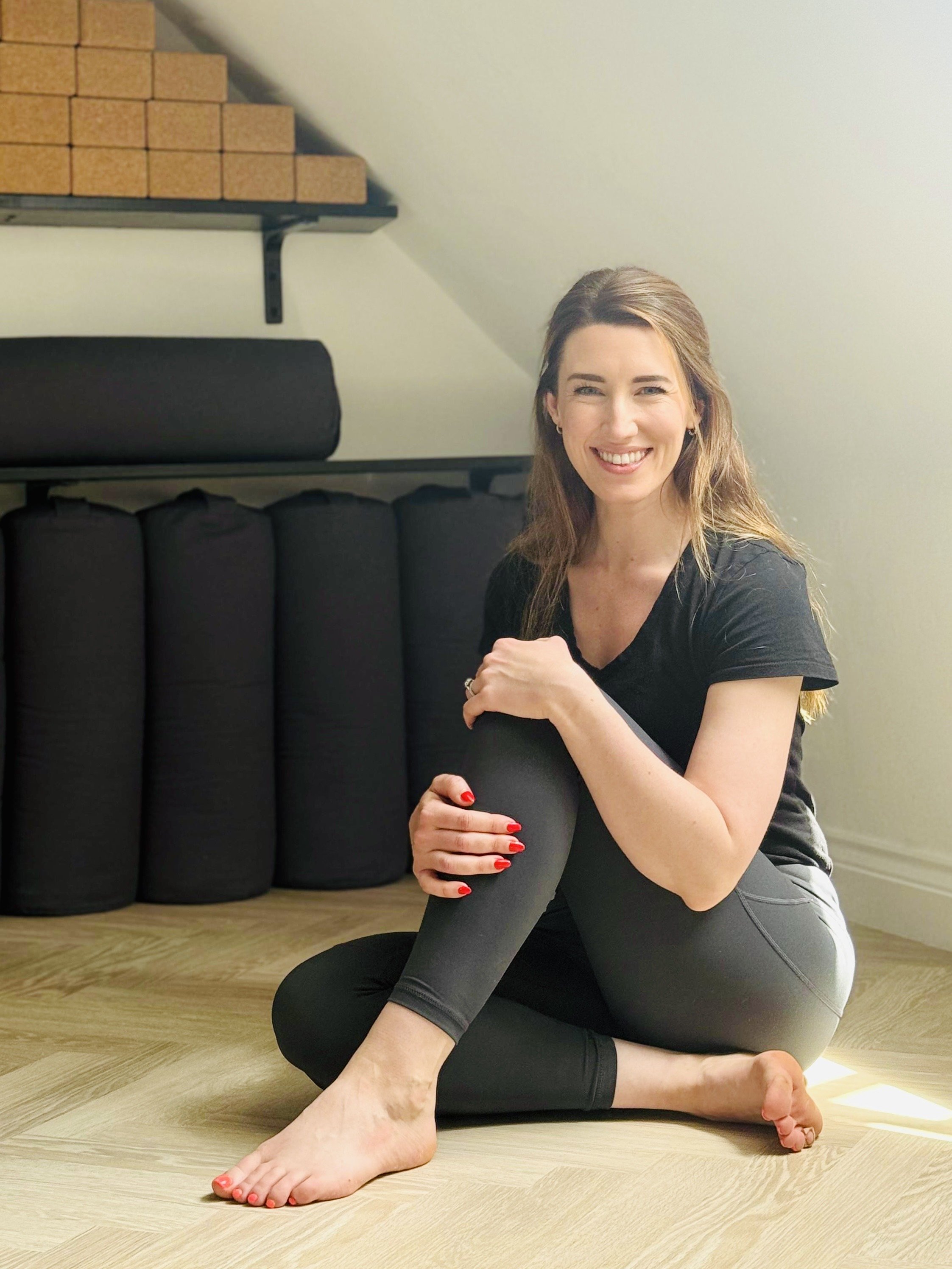 A woman sitting on a wooden floor, smiling, wearing black athletic wear, holding her leg, with a yoga mat and foam rollers in the background.