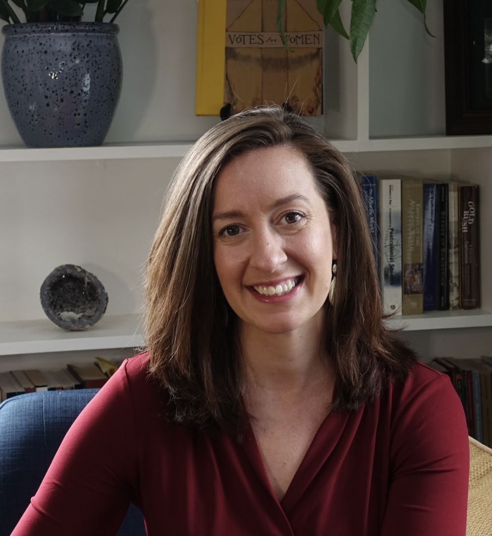 Smiling image of Elena K. Abbott in red blouse, sitting in a blue chair with shelves behind her including a book titled "Votes for Women," as well as a geode, history books, and houseplants
