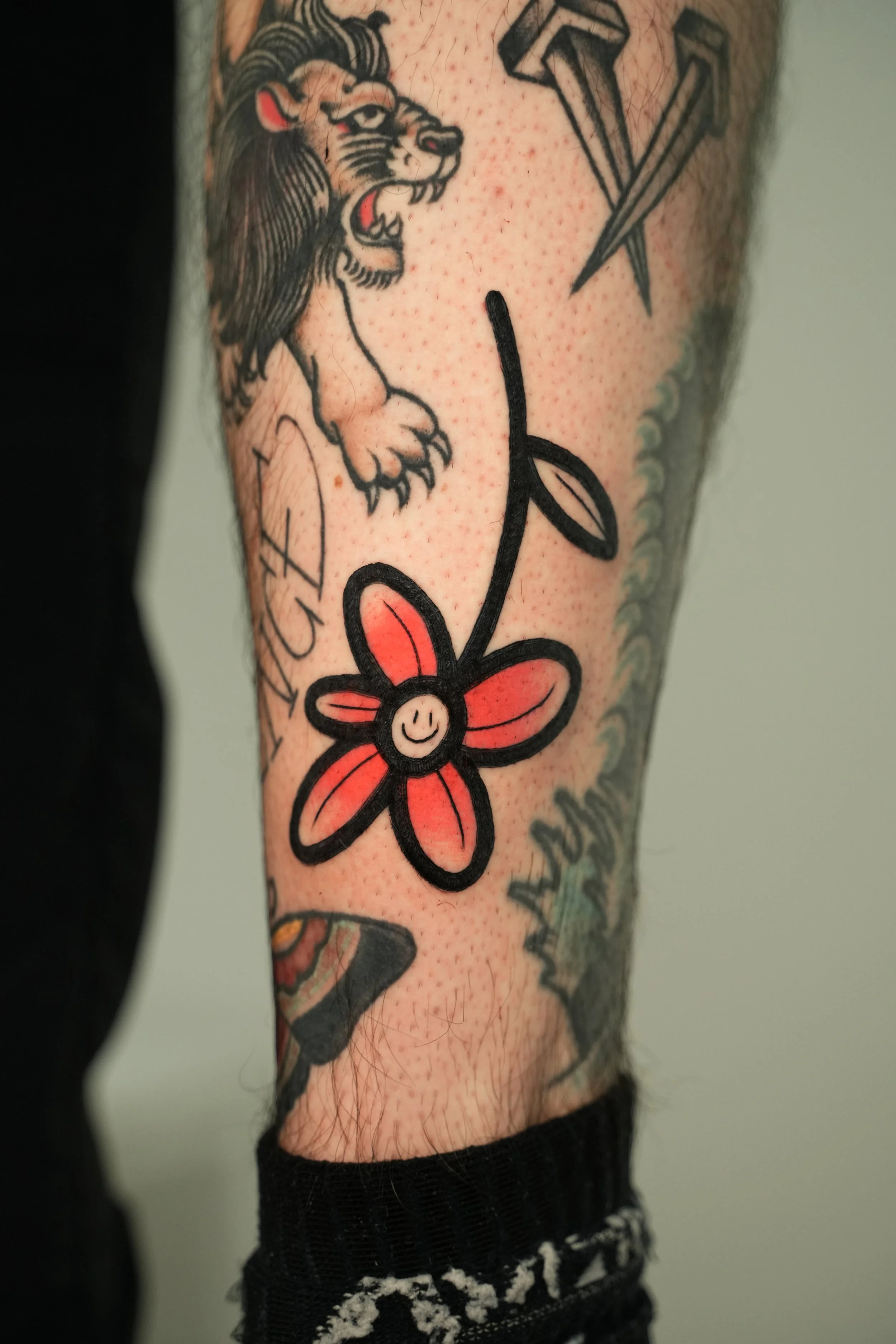 Tattoo of a smiling flower with red petals and black outline on a person's arm, near a traditional style lion tattoo and a pair of nails. The tattoo is colorful with black outlines.