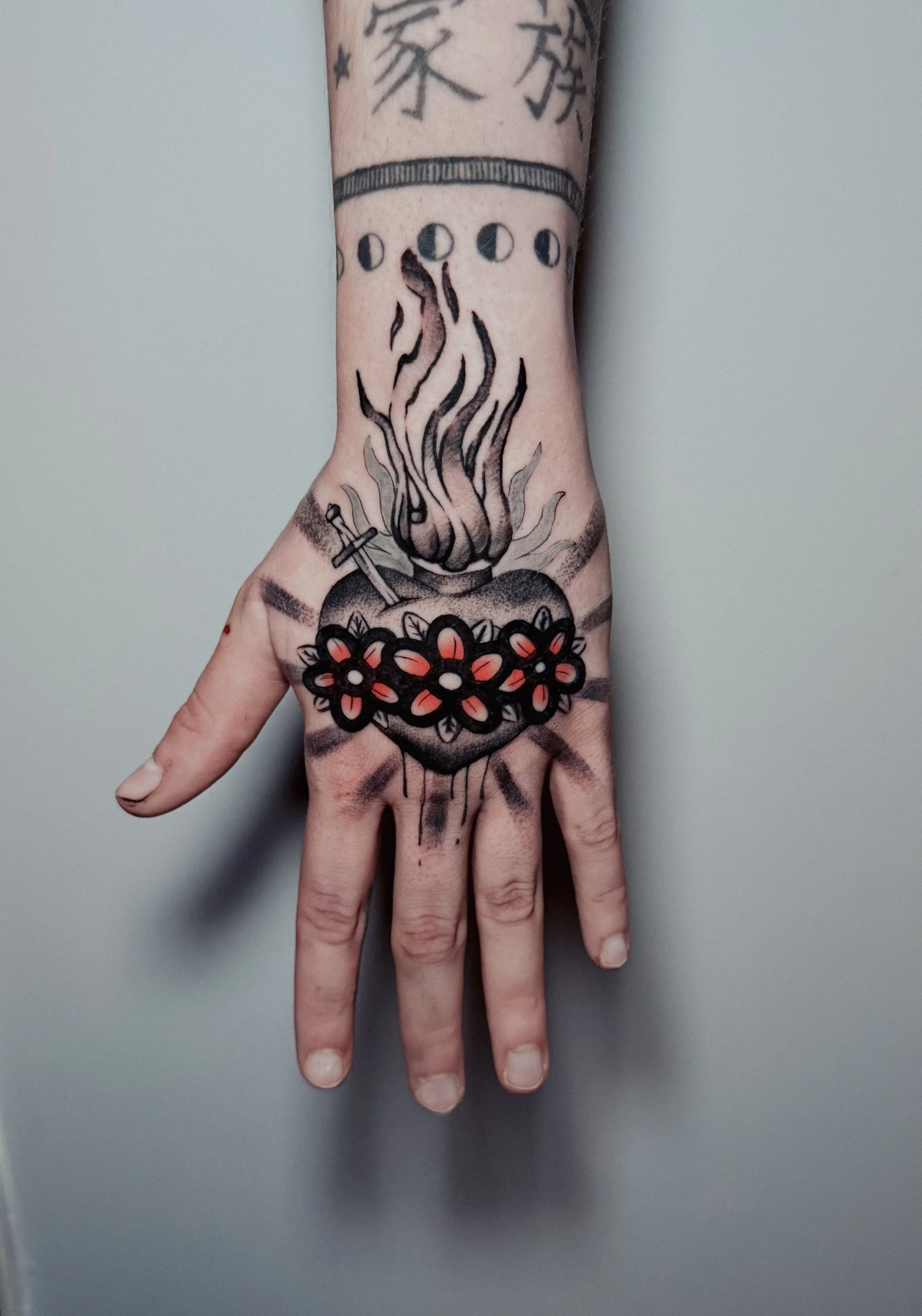 Tattoo of a lit flaming cauldron with a sword, surrounded by flowers on the back of a person's hand and wrist.