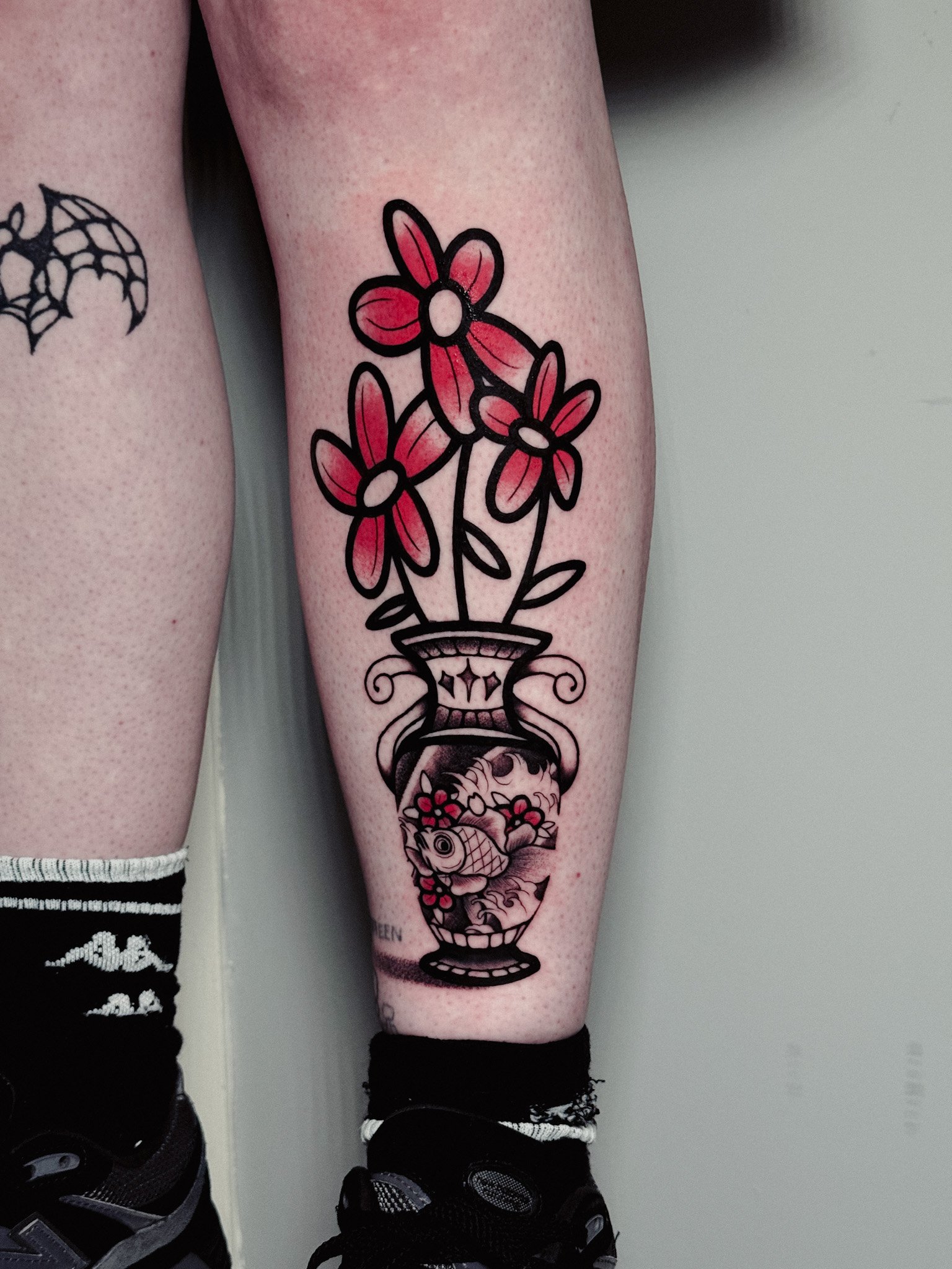 Tattoo of a flower vase with pink flowers on a person's leg, with a fish in water and decorative elements inside an ornate vase.