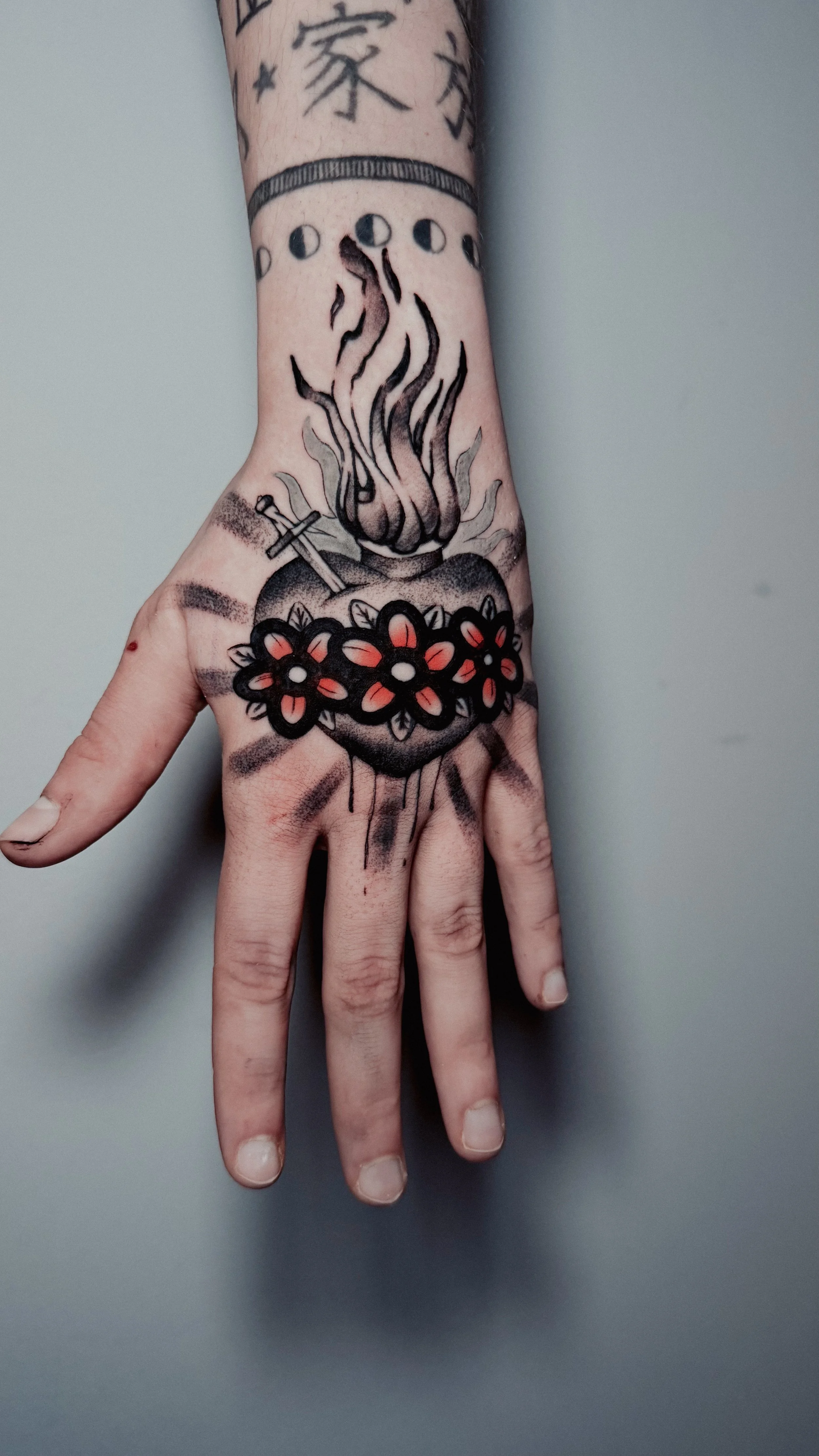 Tattoo on hand featuring a burning lamp with flowers around it, a cross leaning against the lamp, and a moon phase design above.