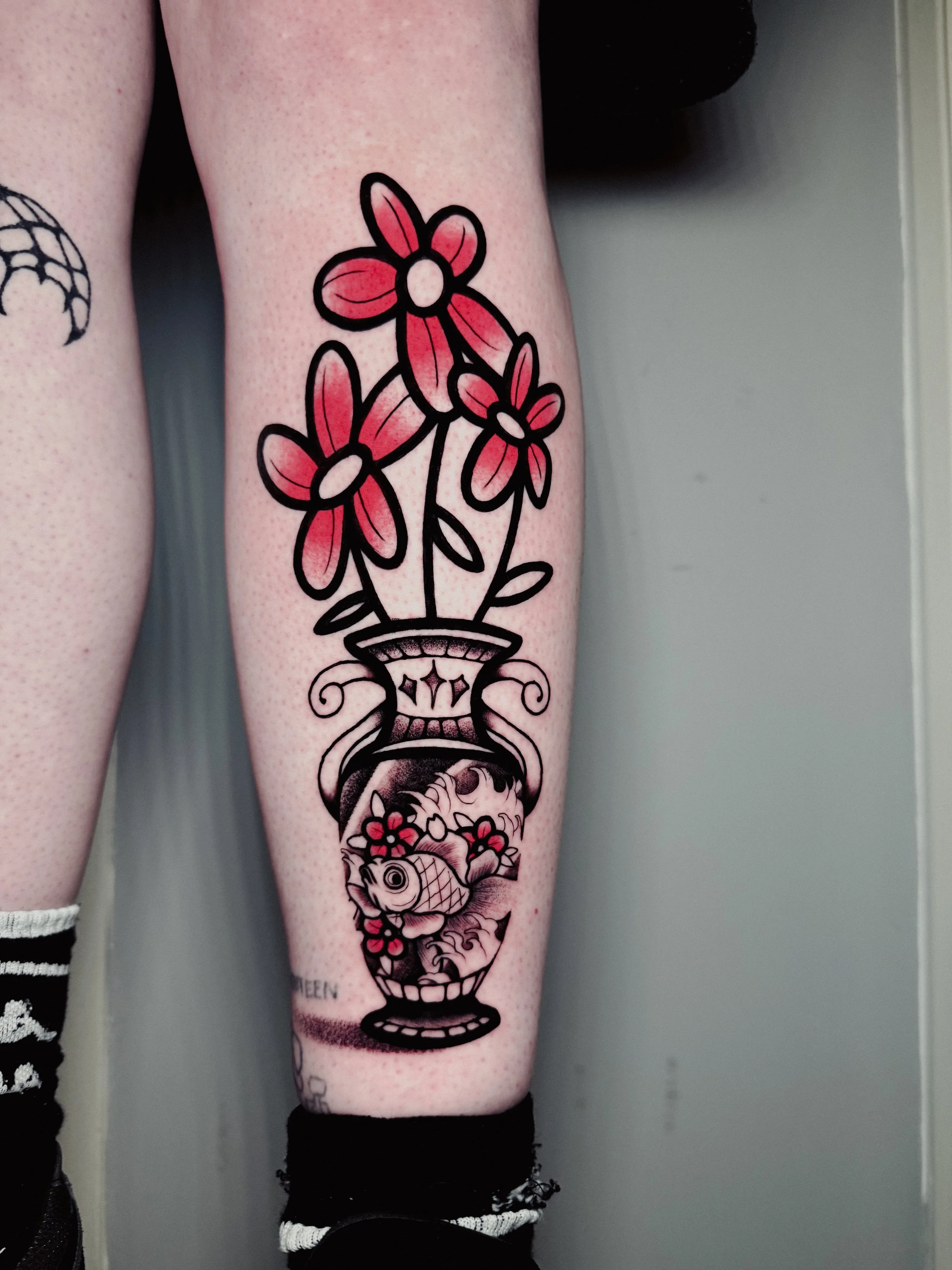 Tattoo of pink flowers in a vase with a fish at the bottom on a person's lower leg.