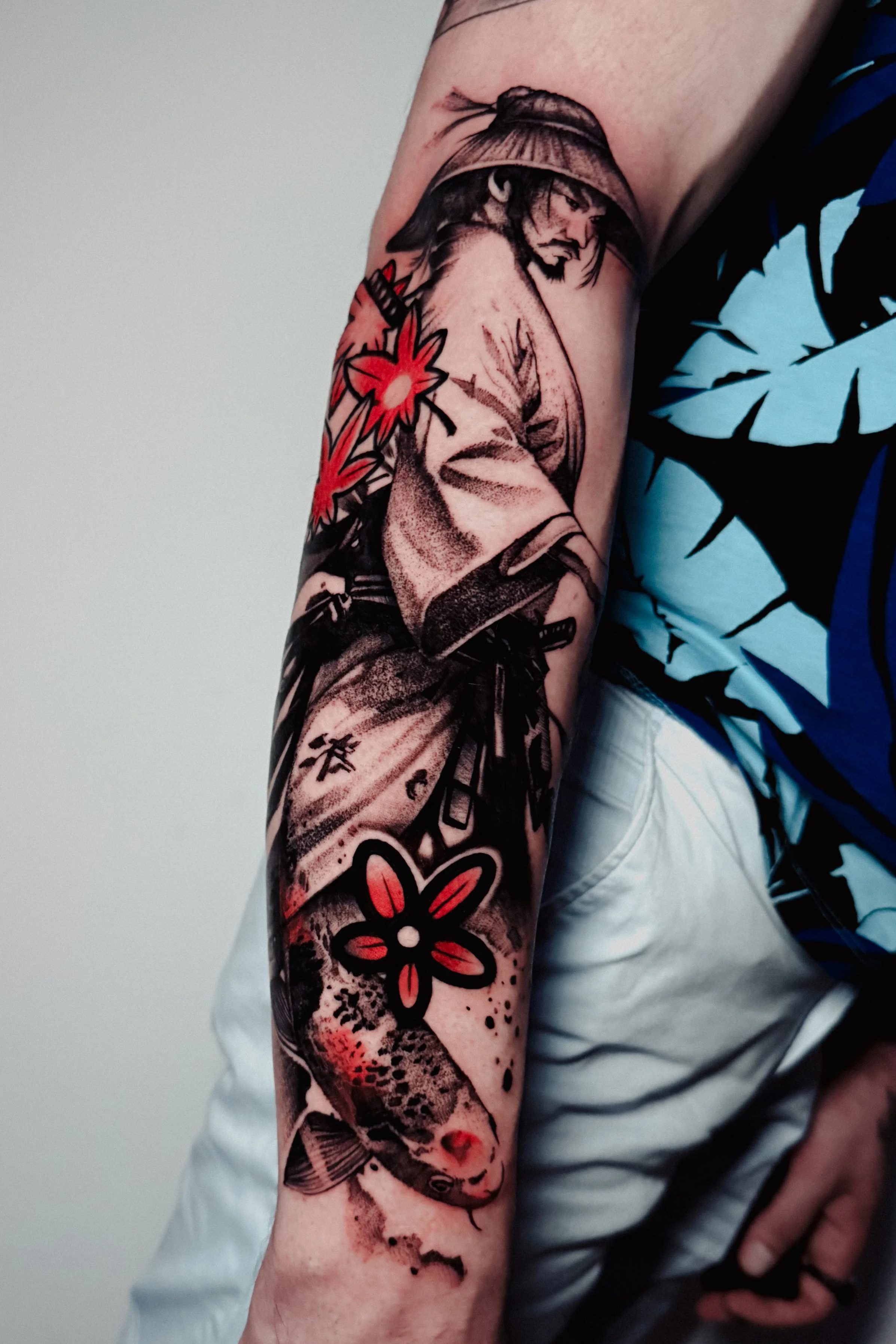 Tattoo of a surrealist samurai man with a hat, a red flower, and a fish in black and red ink on a person's forearm.