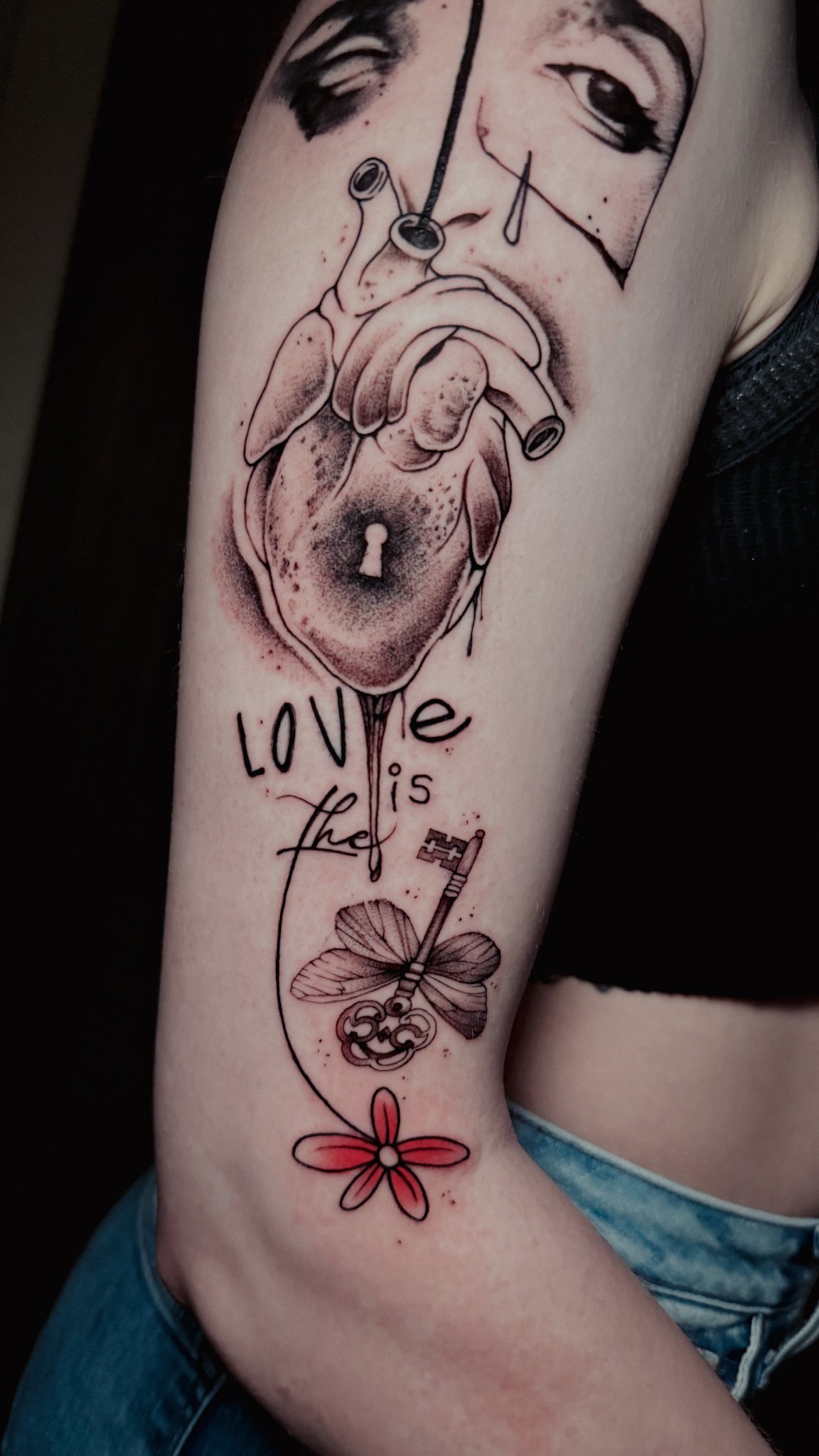Tattoo of a realistic heart with a keyhole, above the phrase "love is the" and a flower, all on a person's arm.