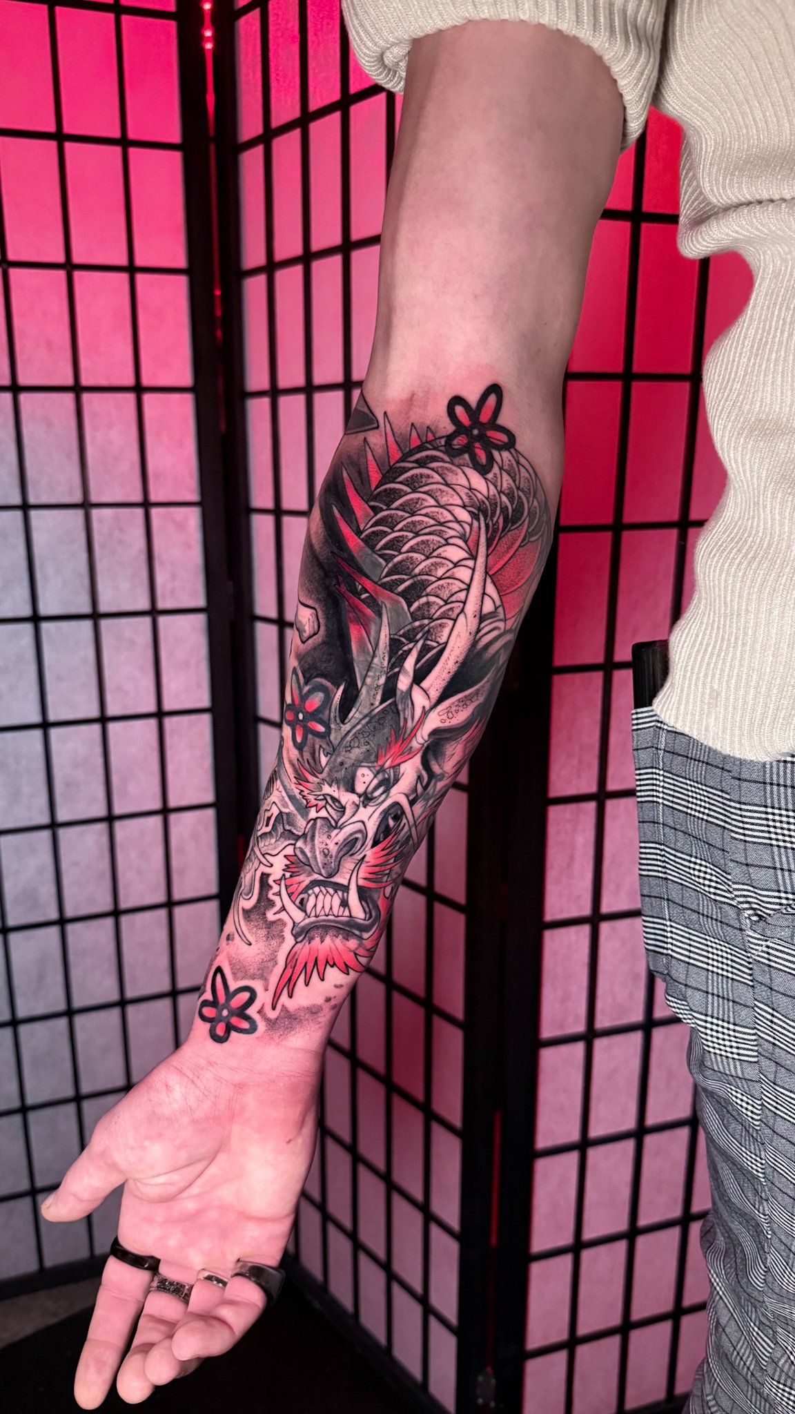 A colorful tattoo of a fierce dragon's face with sharp teeth and red accents on a person's arm, against a pink and black grid background.