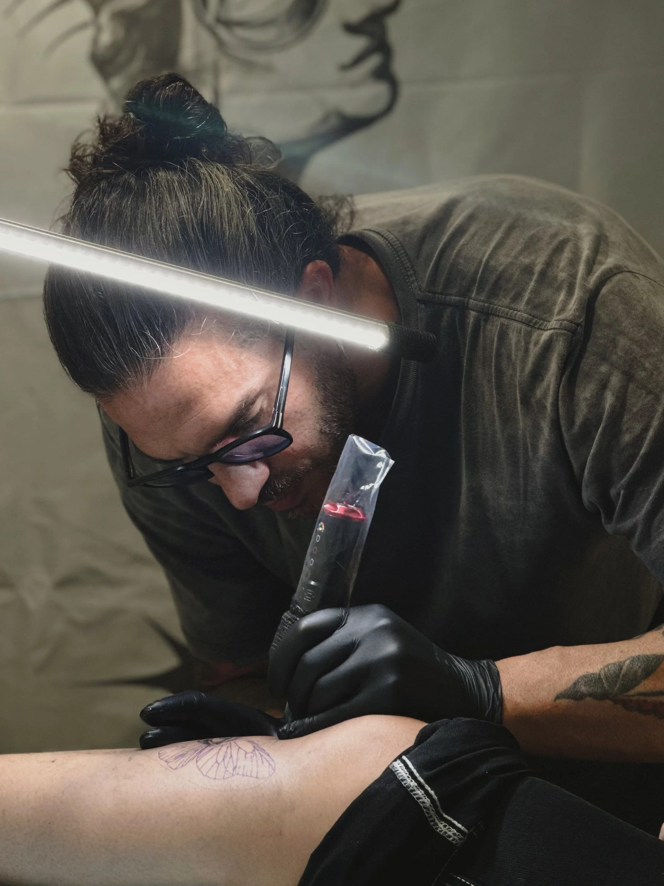 Tattoo artist with black gloves and glasses tattooing a butterfly design on a person's arm.