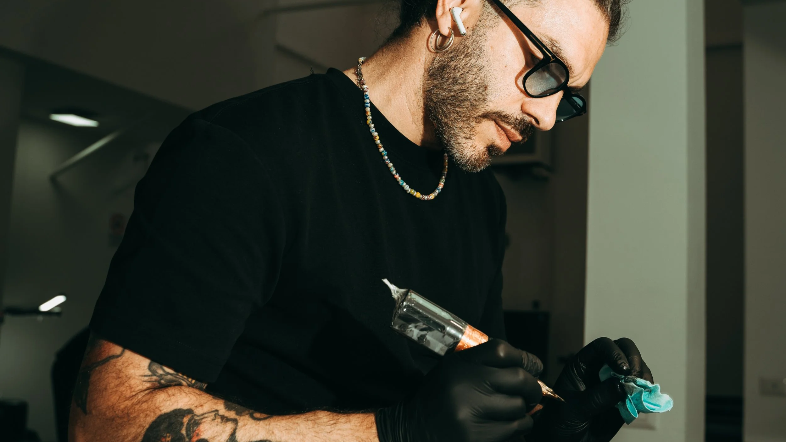 A person with tattoos, tattoos, glasses, and earbuds tattoos is tattooing a design onto a small piece of fabric. The person is wearing black gloves and holding a tattoo machine.