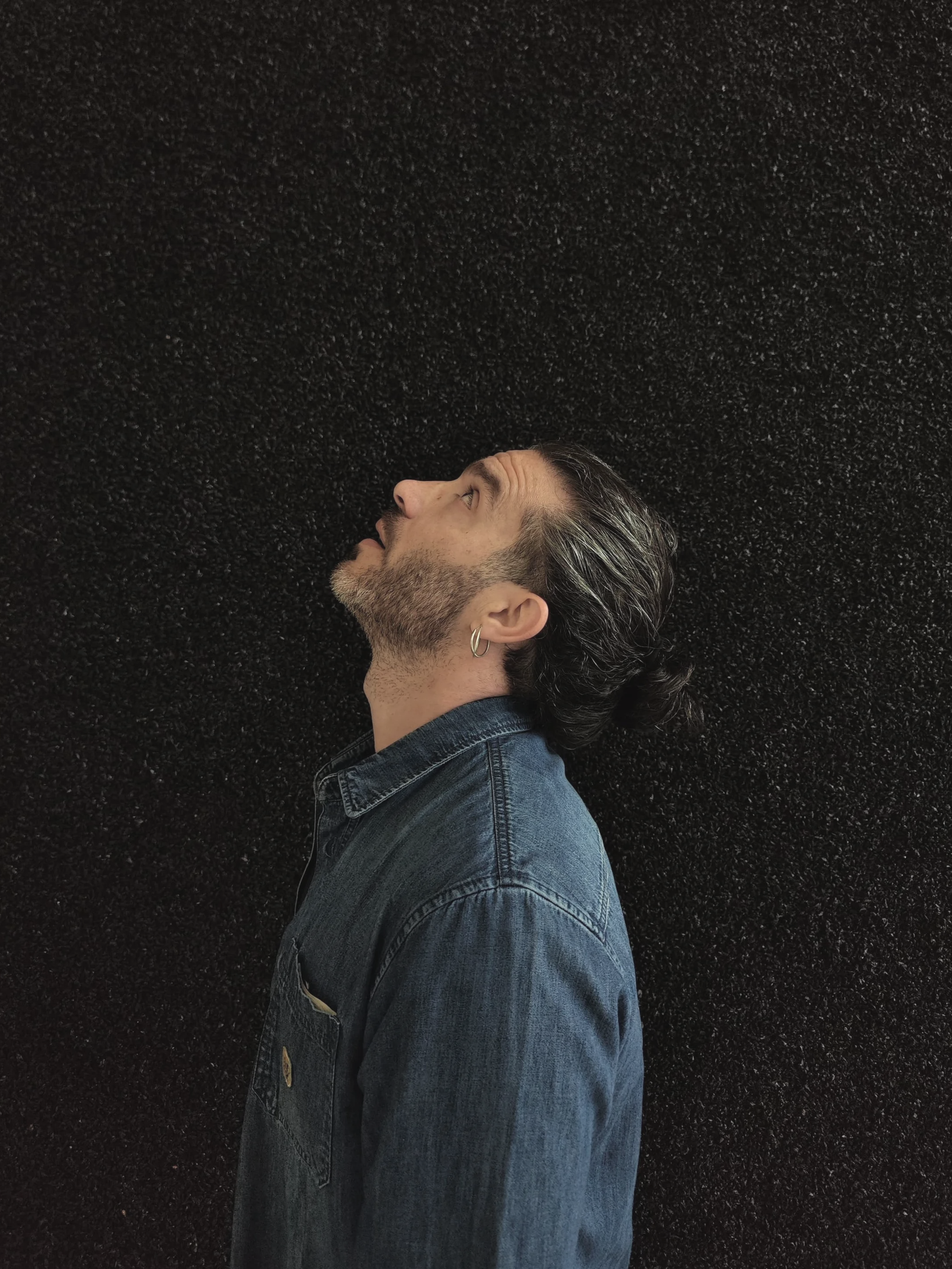 Side profile of a man with dark hair and a beard, wearing a denim jacket, looking upward against a black textured background.