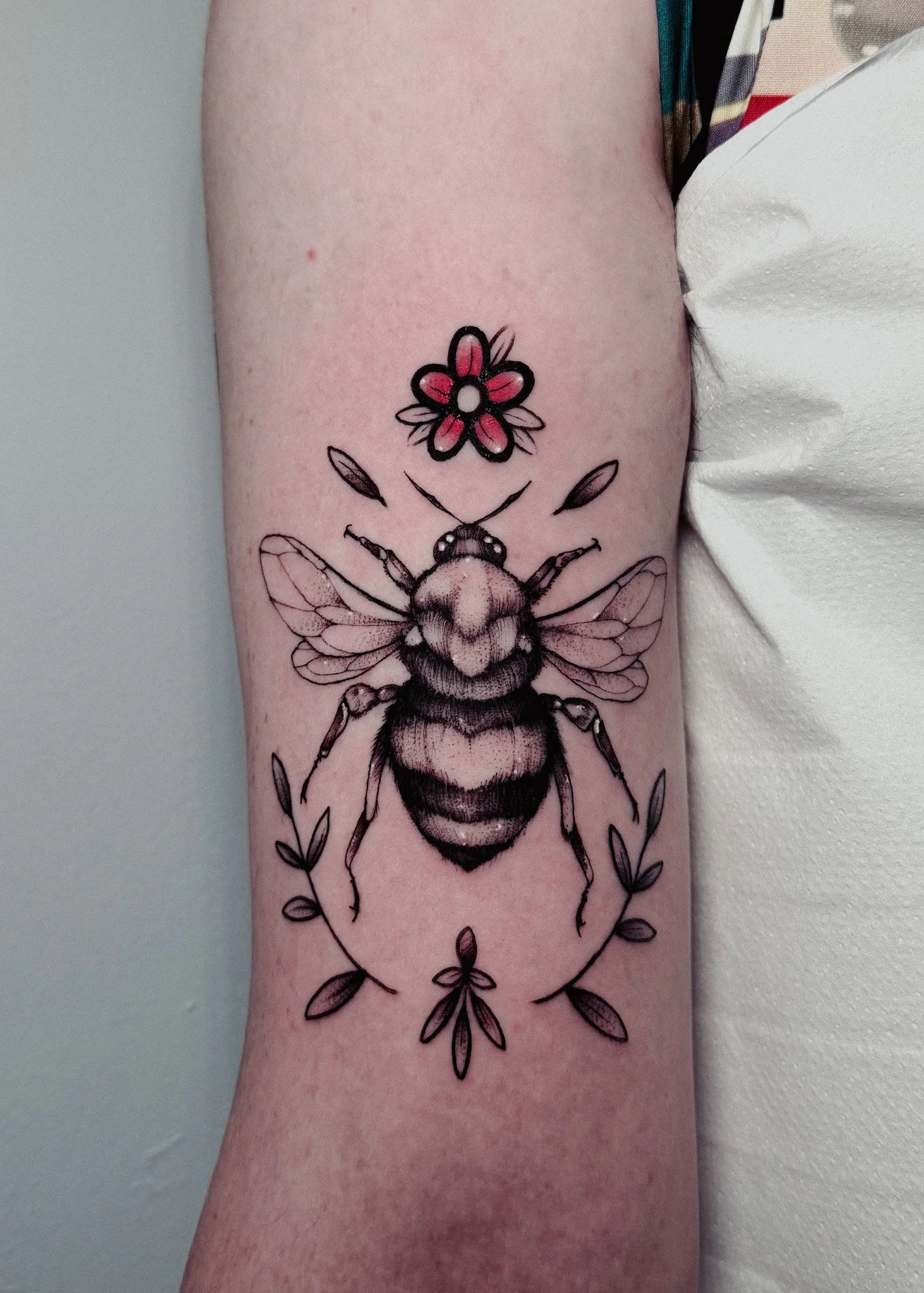 Tattoo of a bee surrounded by small flowers on a person's upper arm.
