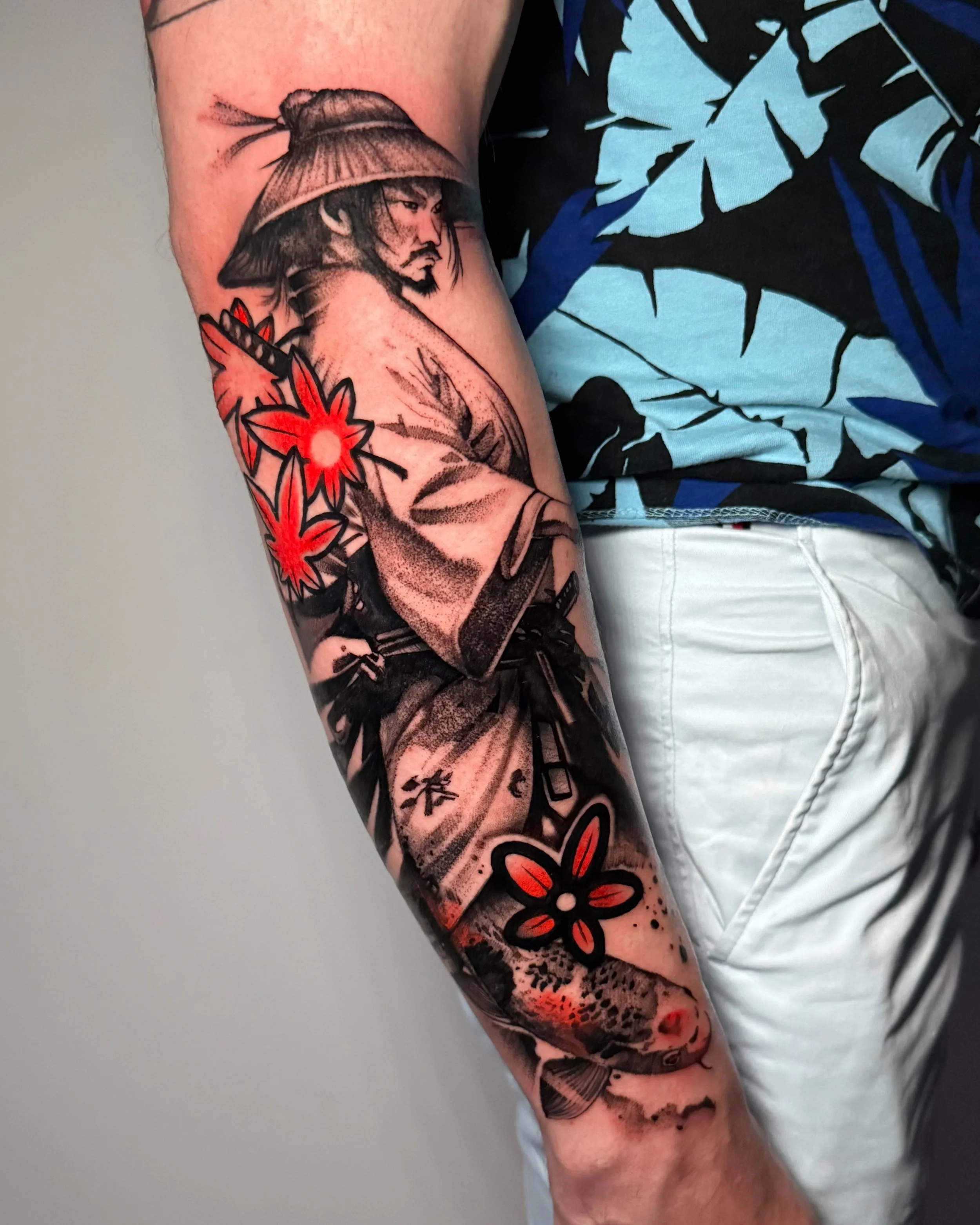 Tattoo of a samurai man with a traditional hat, surrounded by red and black flowers, on a person's arm.