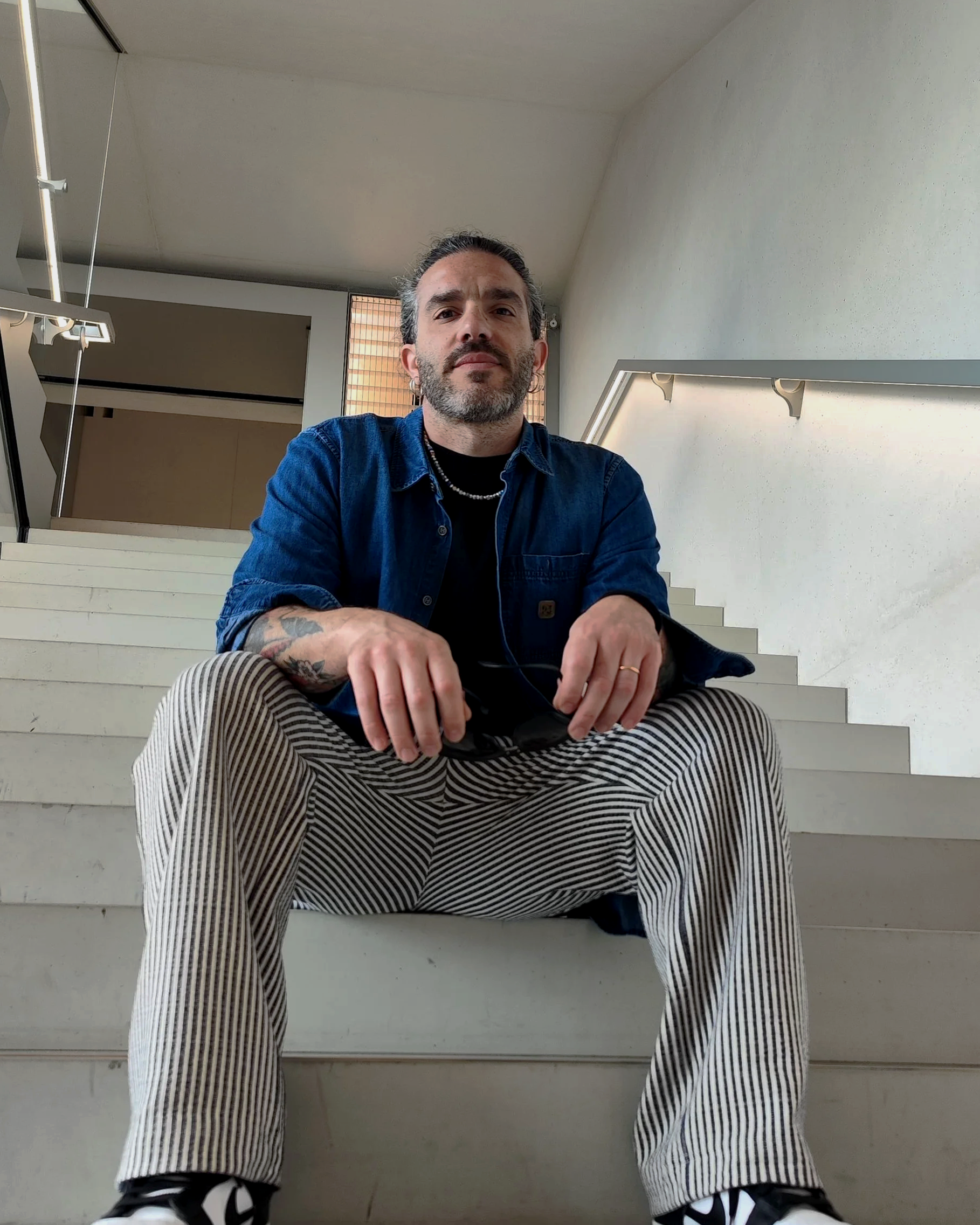 A man sitting on a staircase inside a modern building, wearing a blue denim shirt, striped pants, and holding sunglasses.