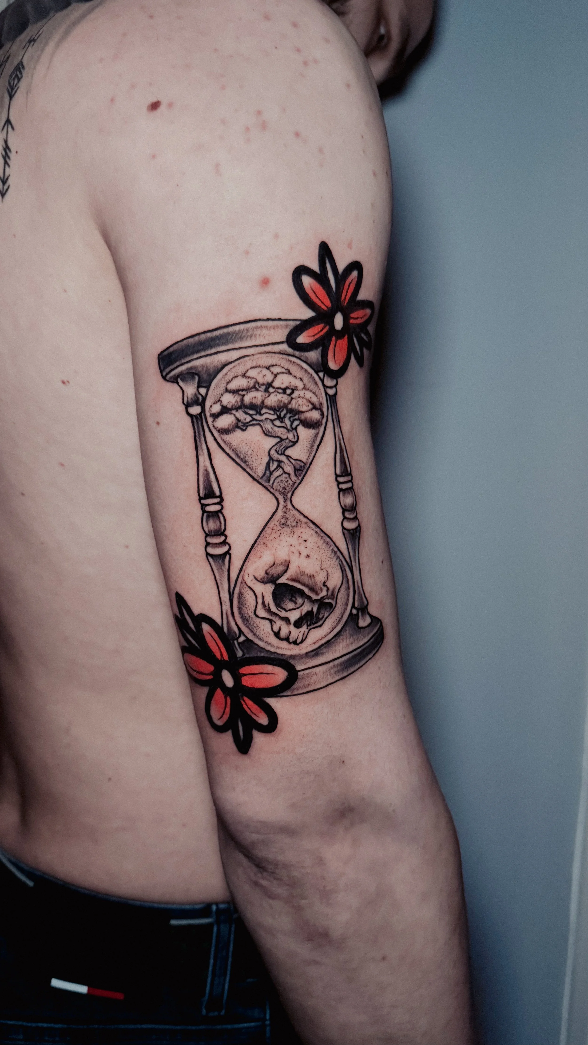 Tattoo of an hourglass with a tree and skull inside, accompanied by red and black flowers on the upper and lower sides, on a person's upper arm.