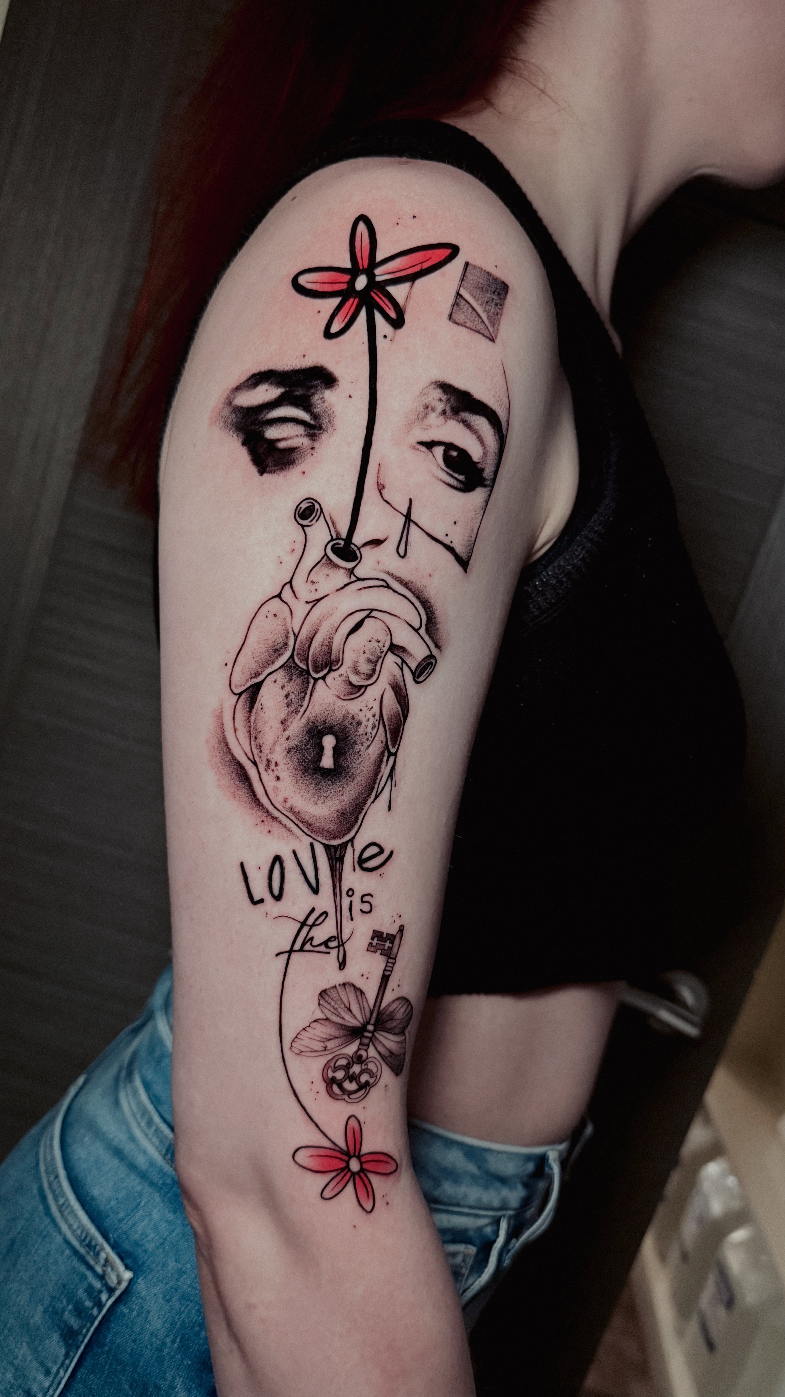 Tattoo of a human heart, with a face and a flower growing from it, along with the phrase "Love is the" and a key with a four-leaf clover on it, and small flowers.