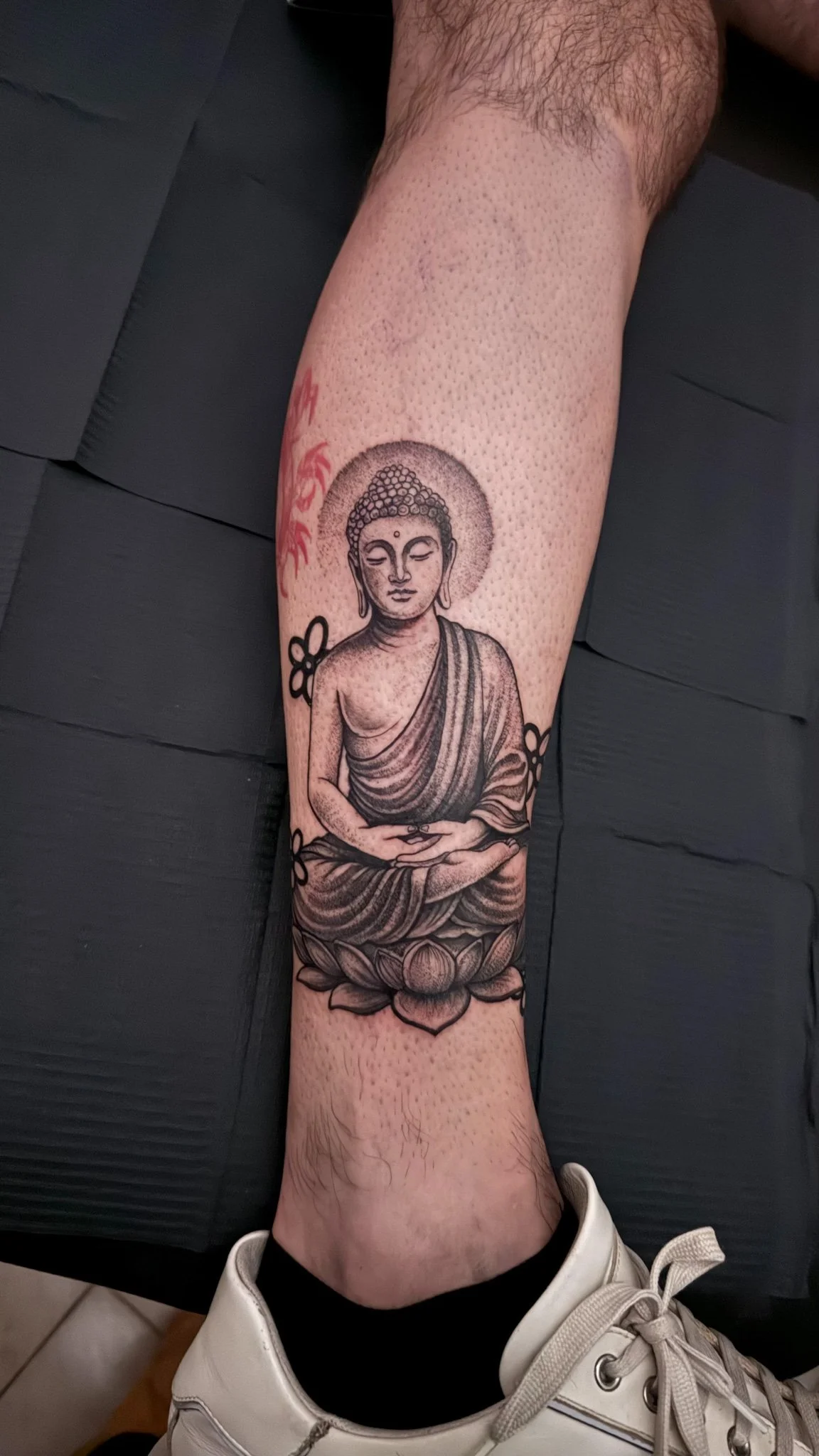 Tattoo of a seated Buddha on a person's lower leg, with a lotus flower base and decorative flowers around it.