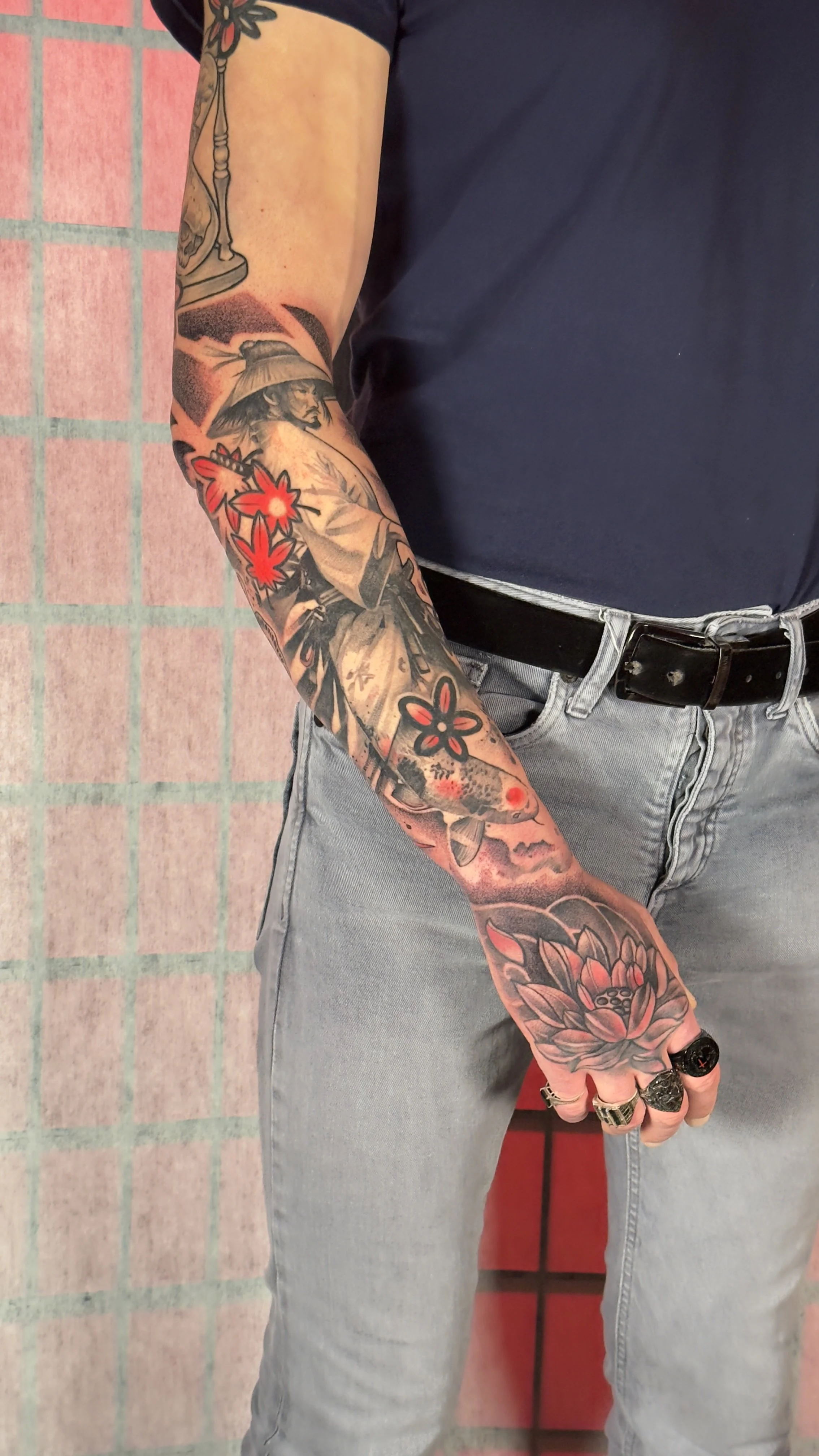 A person with a sleeve tattoo featuring various designs including a samurai character, a koi fish, lotus flowers, and abstract elements. The person is wearing a dark t-shirt, light jeans, and rings, with their hand resting near their pocket against a