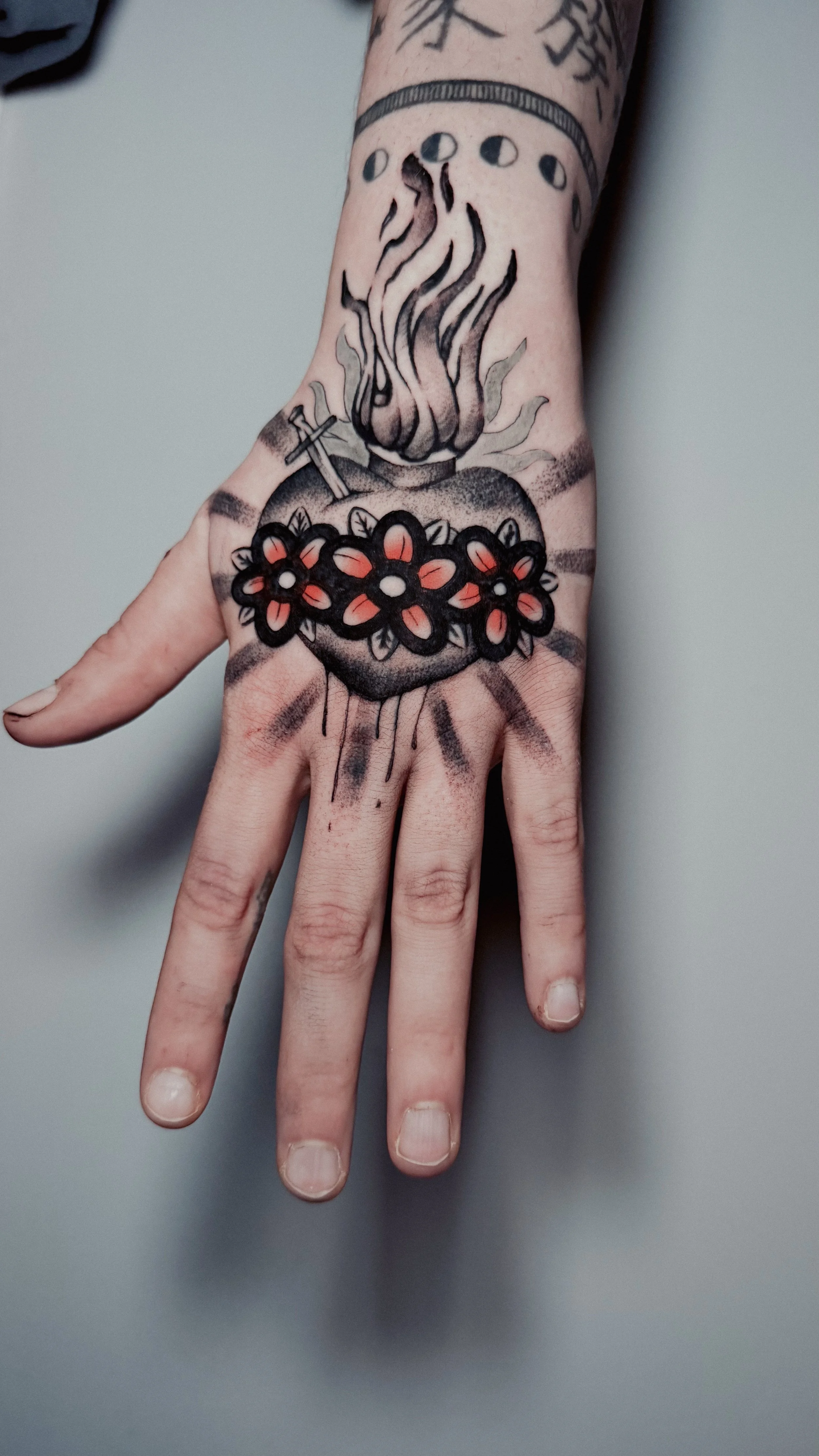 Tattoo of a lit candle on the back of a hand, surrounded by black outlines of flowers and a small cross, with black shading and orange accents.