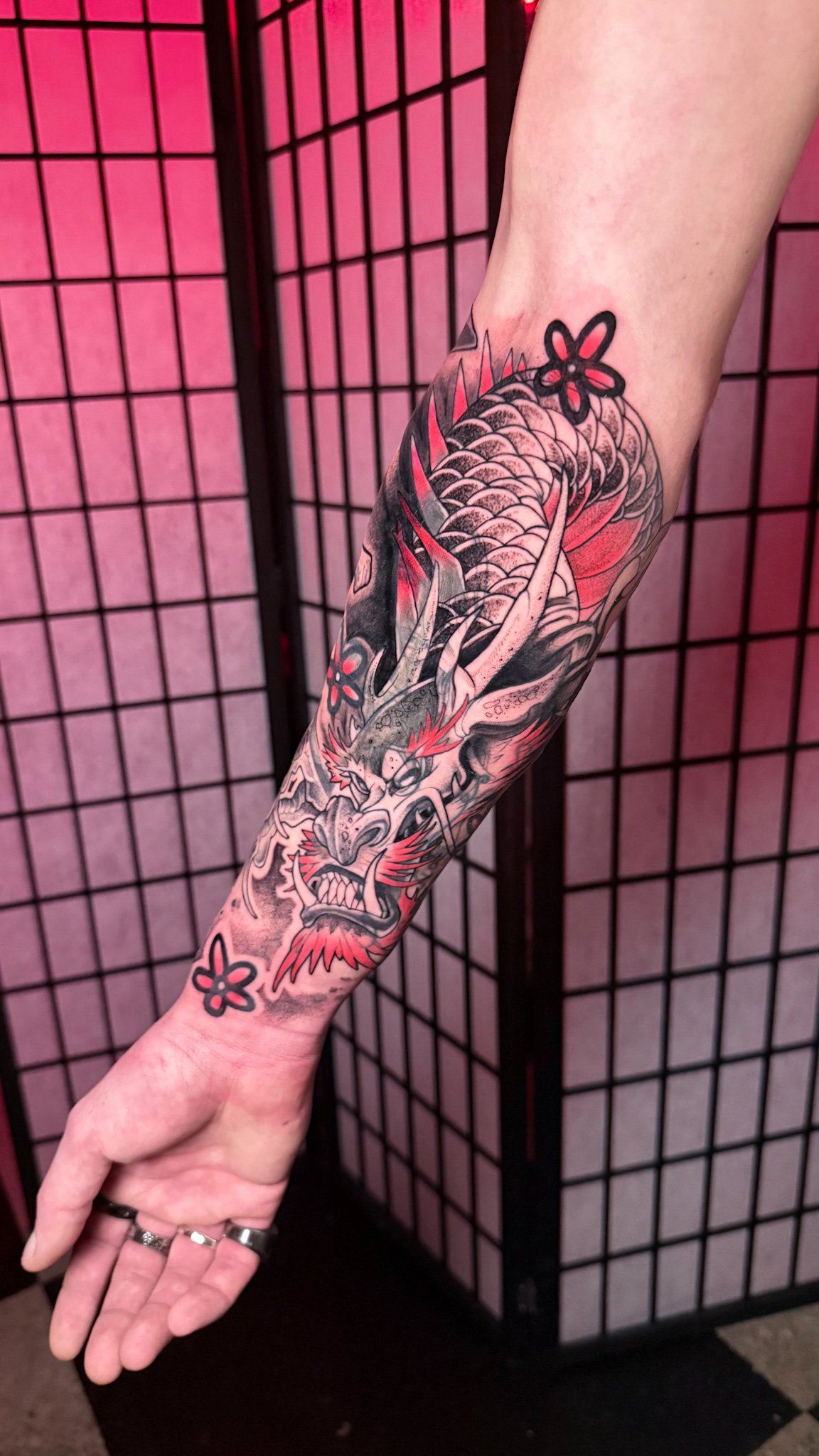 A detailed black and gray tattoo of a snarling dragon with red highlights on a person's forearm, with the dragon's body extending from the wrist to the elbow and a background of a pink and black folding screen.