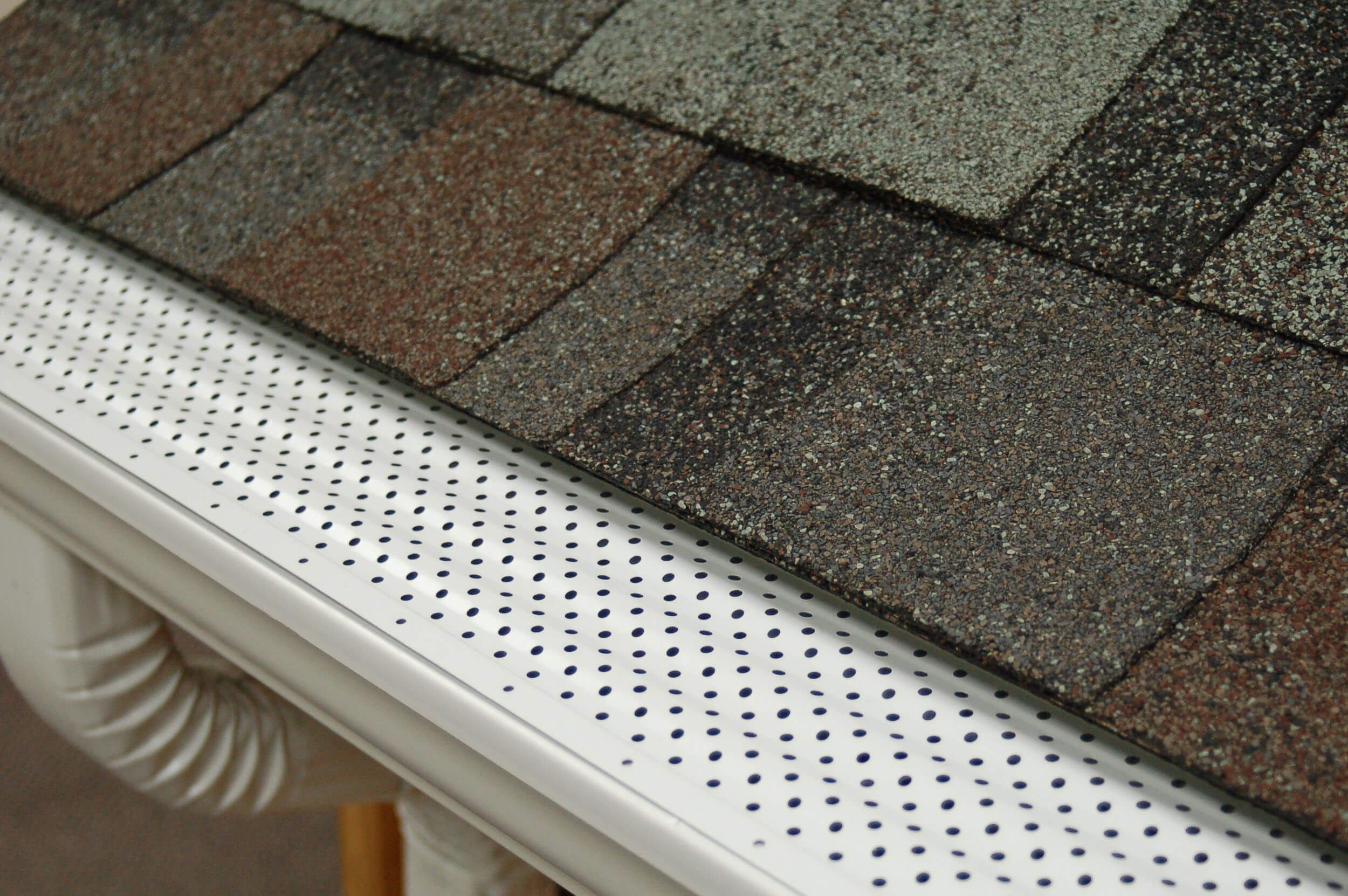 Close-up of asphalt shingles in various shades of brown, gray, and black placed on a white perforated table with a rounded corner.