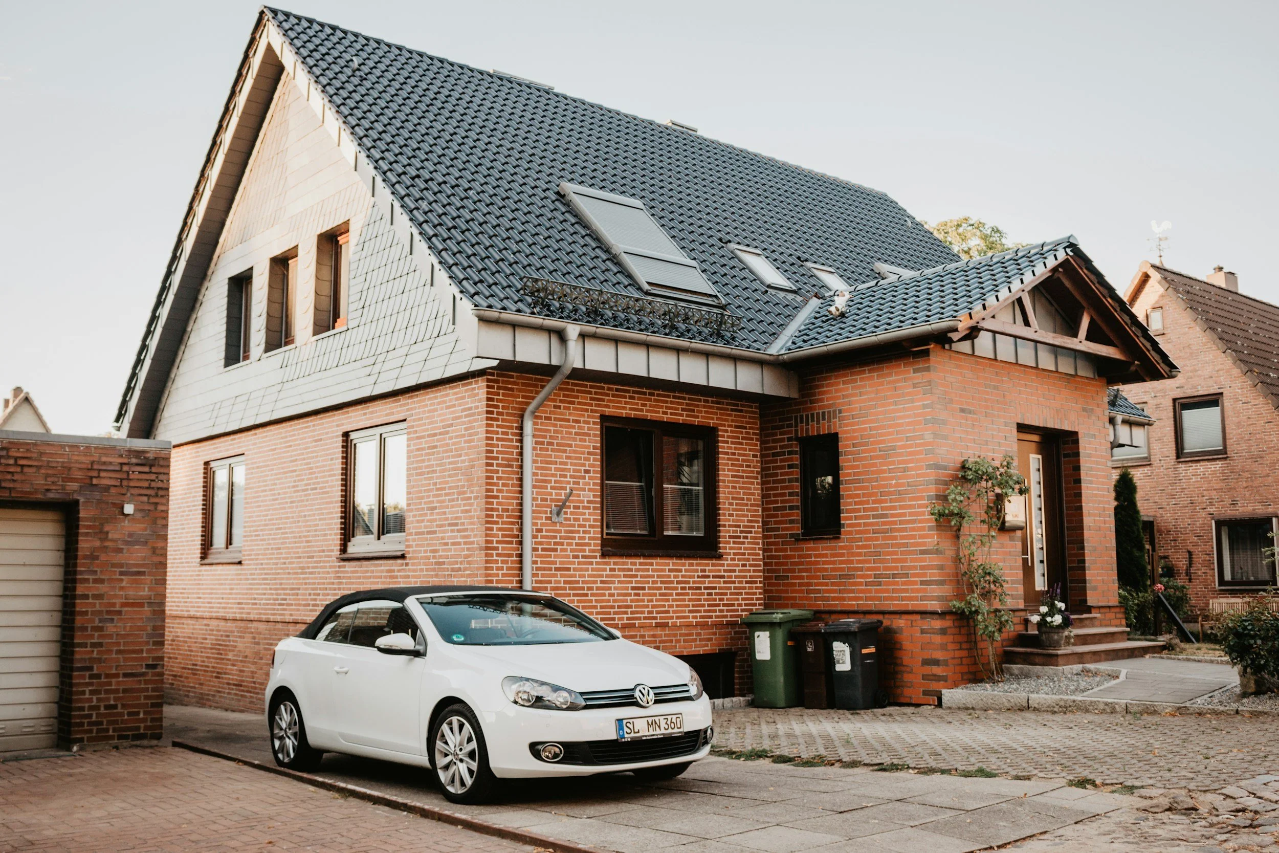 A modern two-story house with a brick exterior, dark gray roof, and several skylights. There is a white Volkswagen car parked in front of the house and a small garden with plants near the entrance.