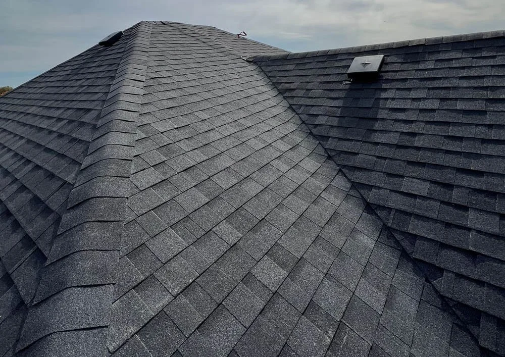 Close-up of a black shingled roof with two ventilation vents under a cloudy sky.