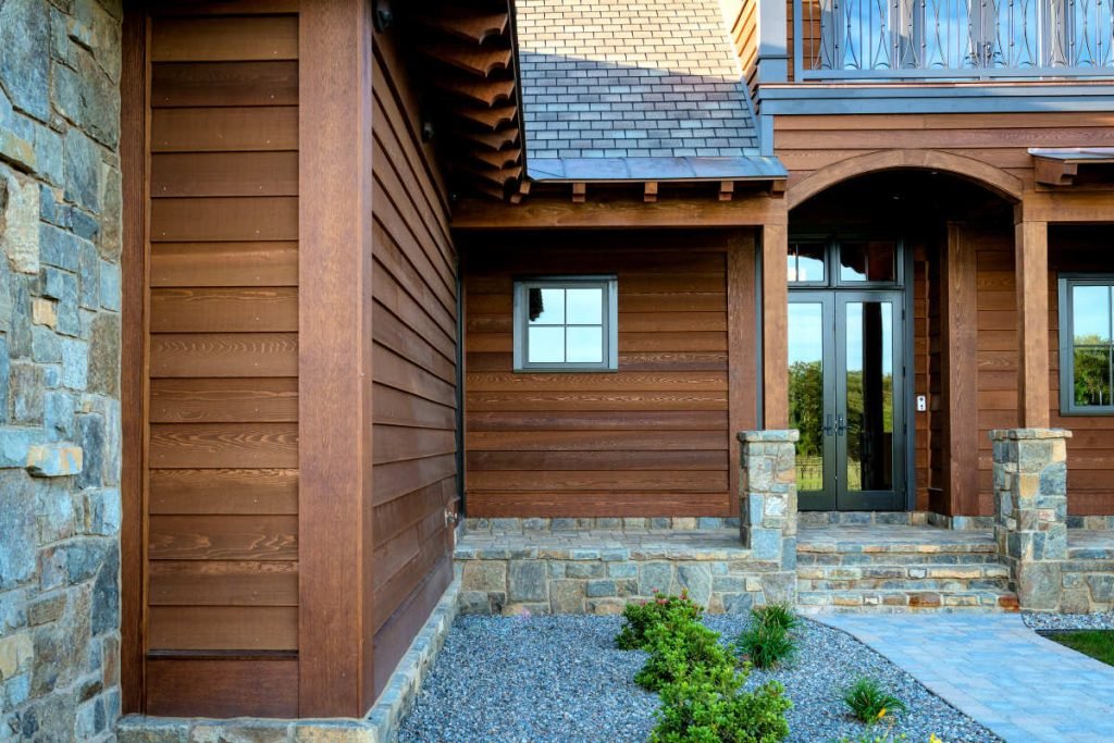 Front entrance of a house with stone steps, wooden siding, glass double doors, small window, and a stone and gravel yard with plants.